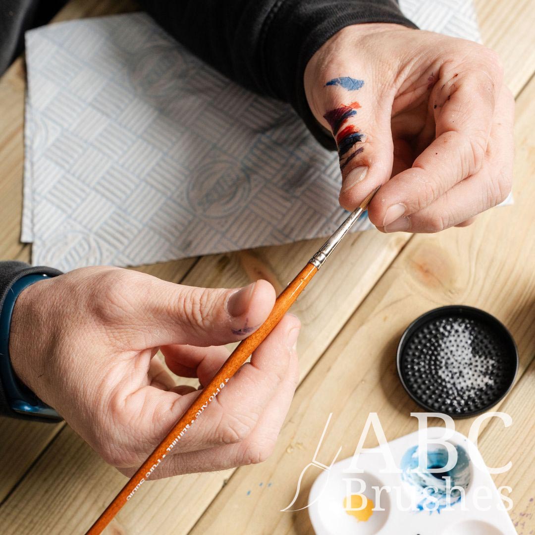 Person reshaping the tip of a Pro Arte Sentinel brush. There is a paint palette, ABC paintbrush cleaner and a paper towel below this. 