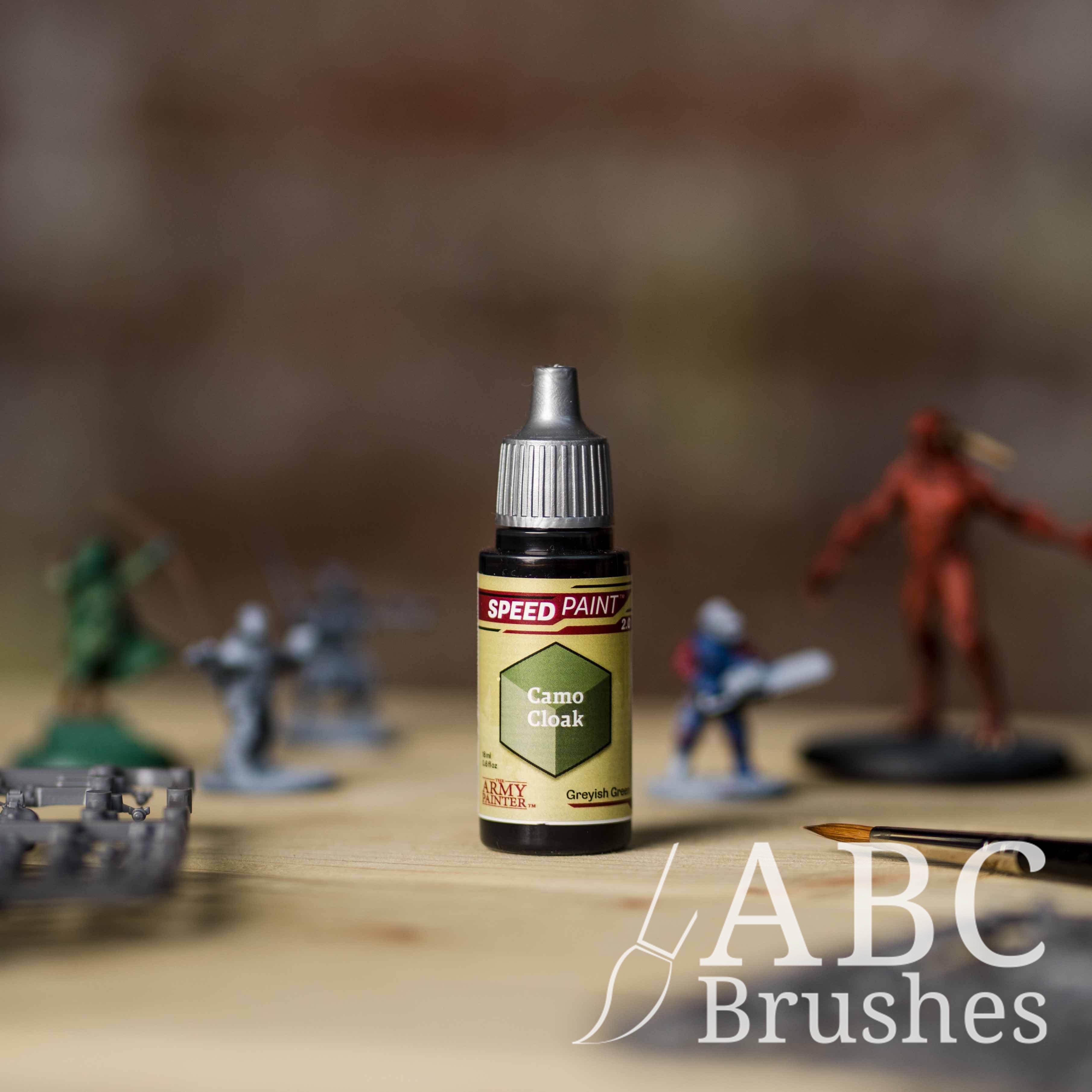 Bottle of the Army Painters Speedpaint in the colour Camo Cloak, with wargaming models and an ABC paintbrush in the background, on a wooden surface.