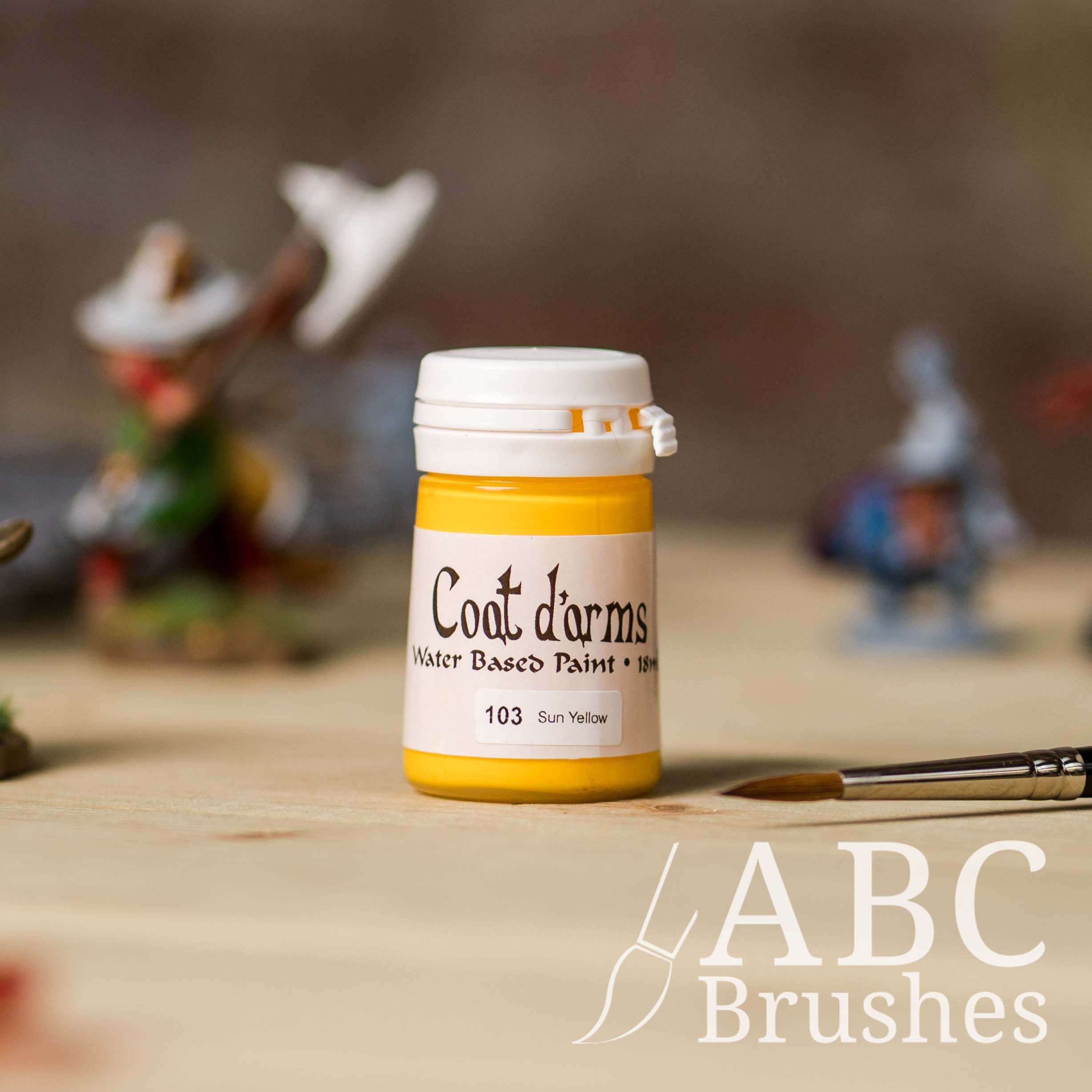Bottle of Coat d'Arms acrylic paint in the colour Sun Yellow, with various wargaming models and an ABC paintbrush in the background, on a wooden surface.