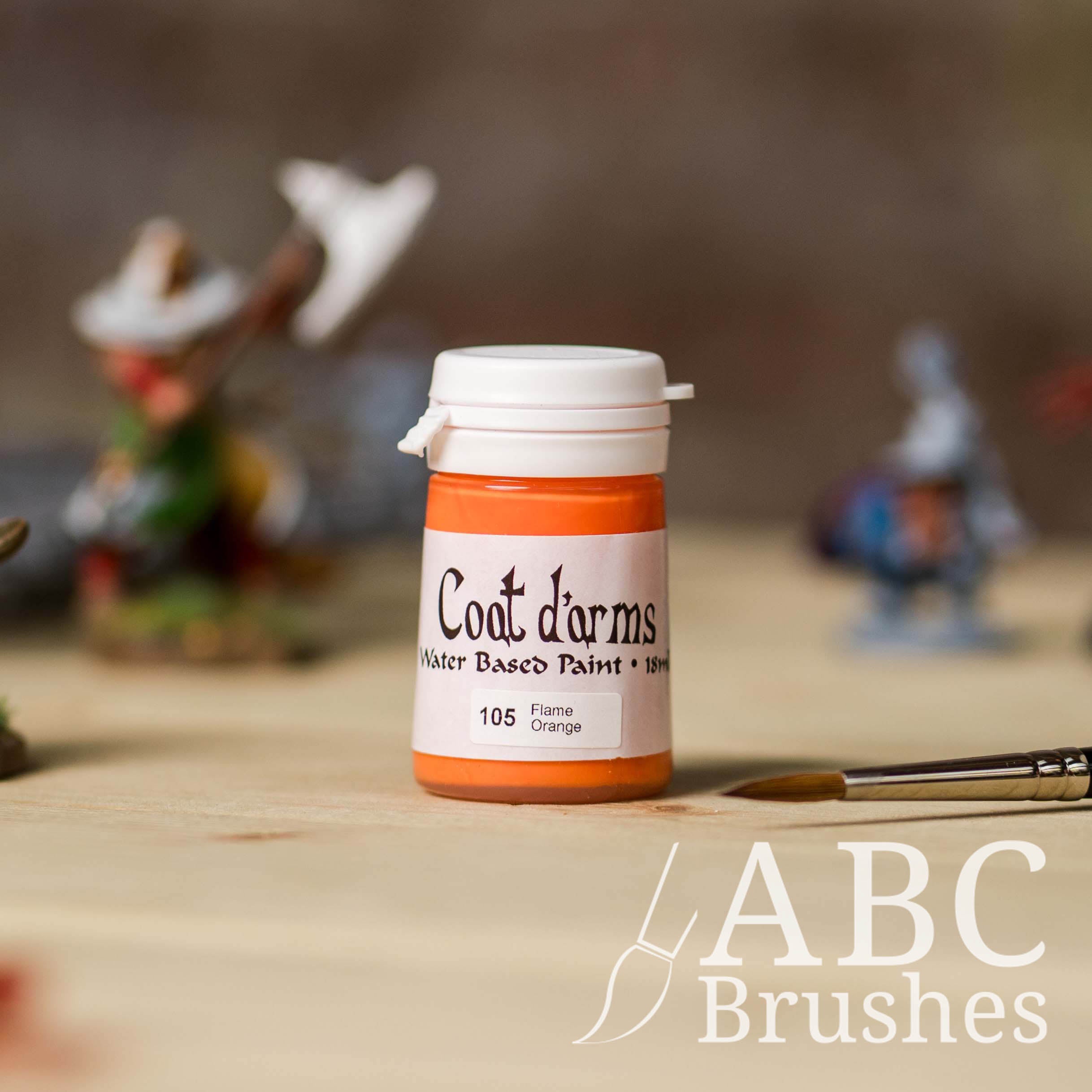 Bottle of Coat d'Arms acrylic paint in the colour Flame Orange, with various wargaming models and an ABC paintbrush in the background, on a wooden surface.