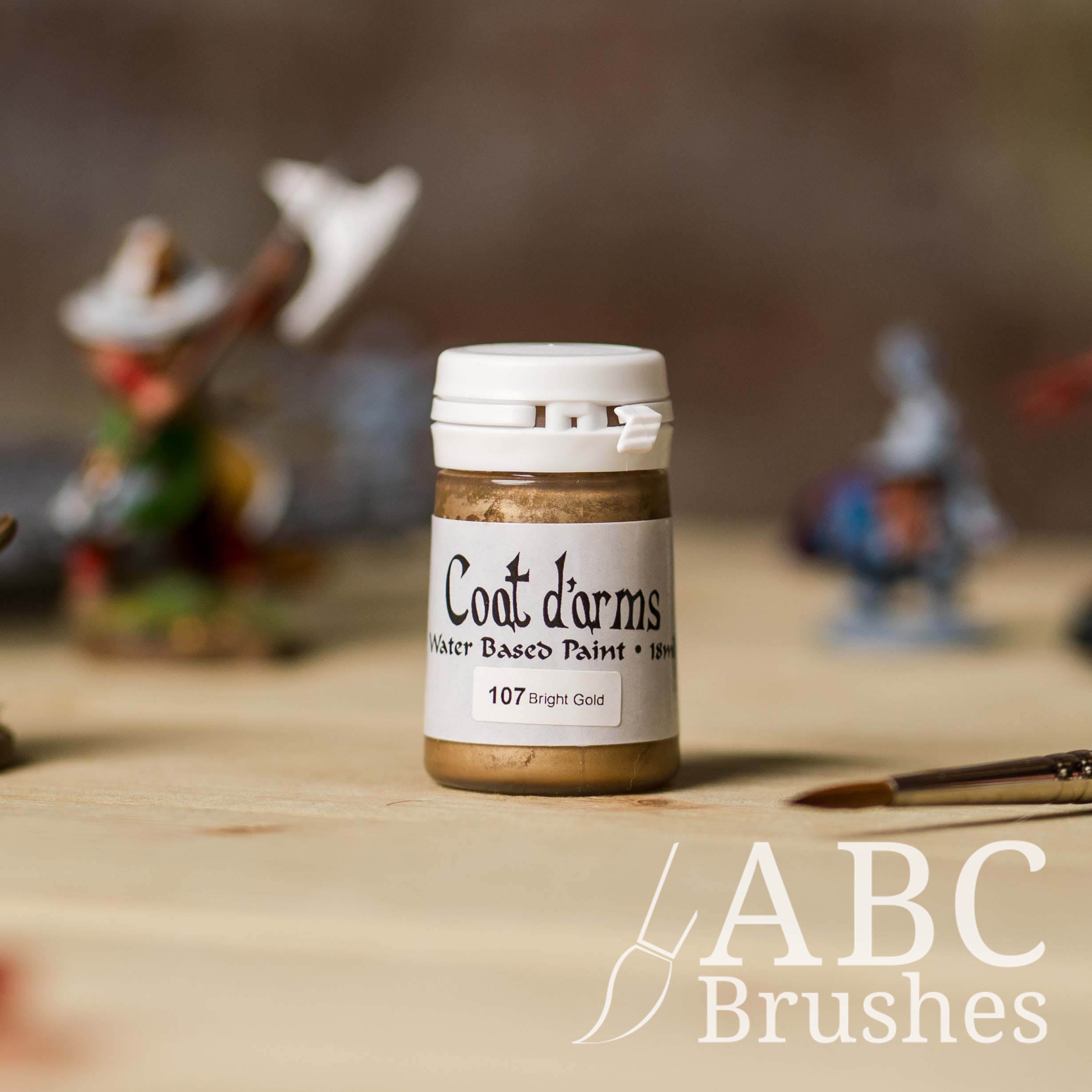 Bottle of Coat d'Arms acrylic paint in the colour Bright Gold, with various wargaming models and an abc paintbrush in the background, on a wooden surface.
