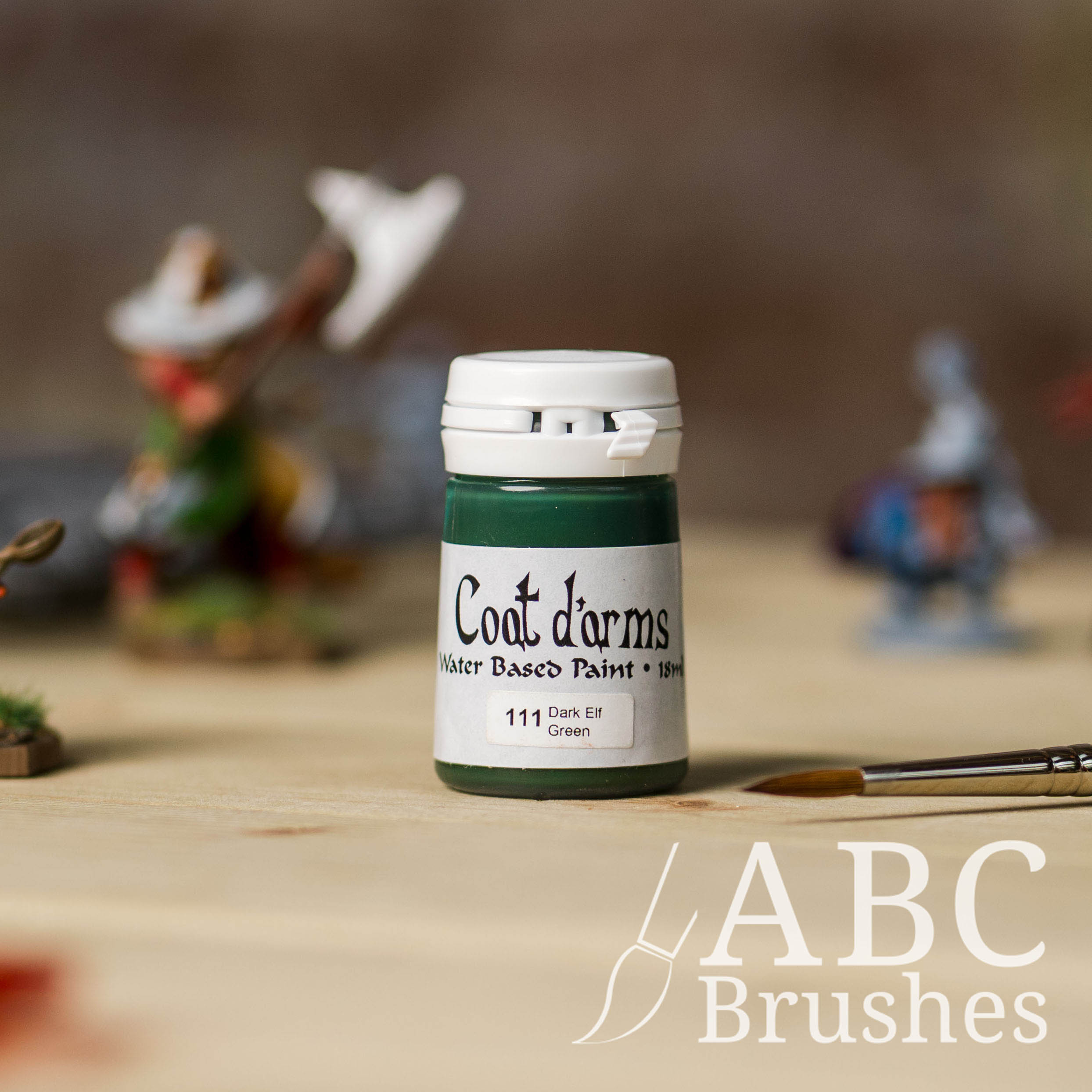 Bottle of Coat d'Arms acrylic paint in the colour Dark Elf Green, with various wargaming models and an abc paintbrush in the background, on a wooden surface.