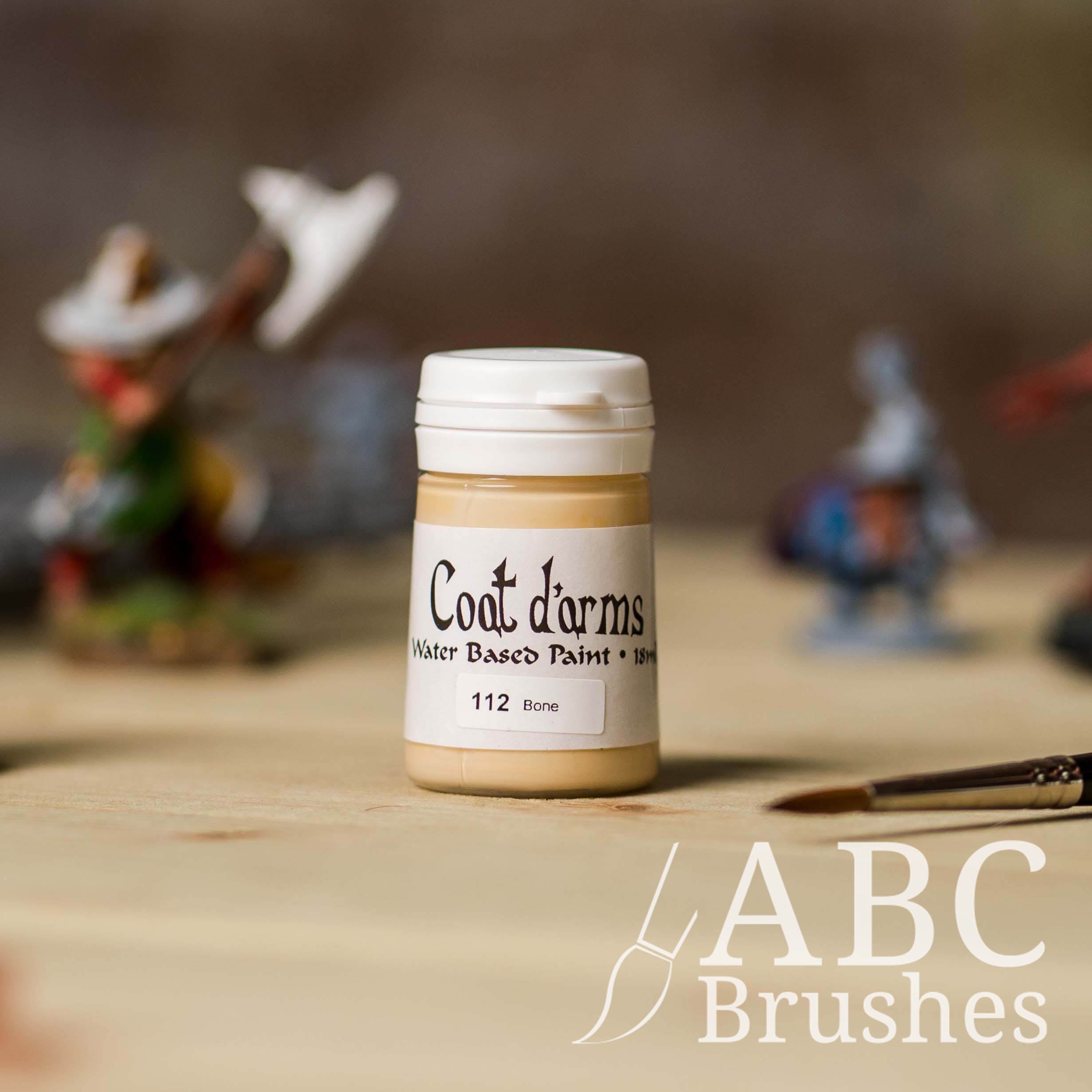 Bottle of Coat d'Arms acrylic paint in the colour Bone, with various wargaming models and an abc paintbrush in the background, on a wooden surface.