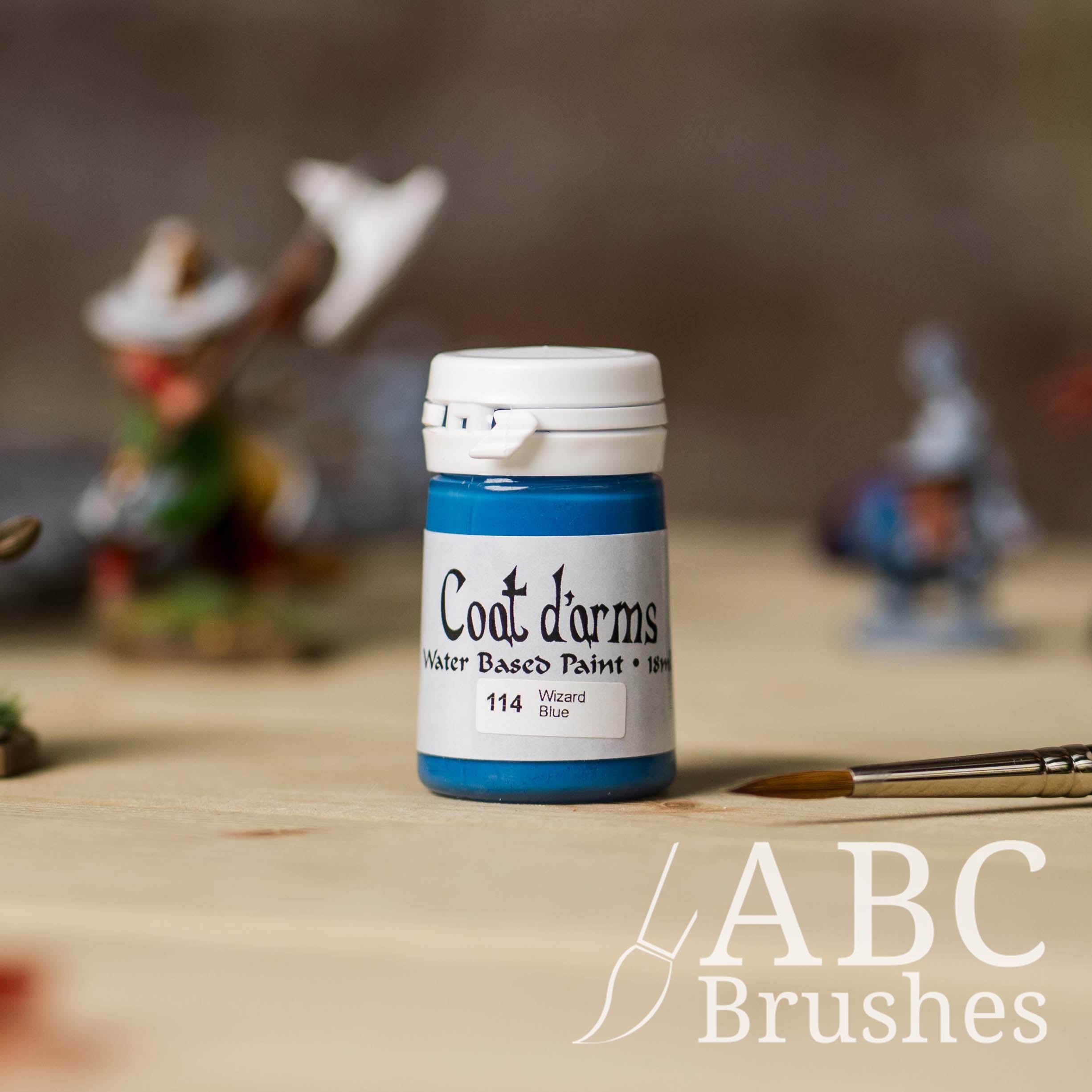 Bottle of Coat d'Arms acrylic paint in the colour Wizard Blue, with various wargaming models and an abc paintbrush in the background, on a wooden surface.