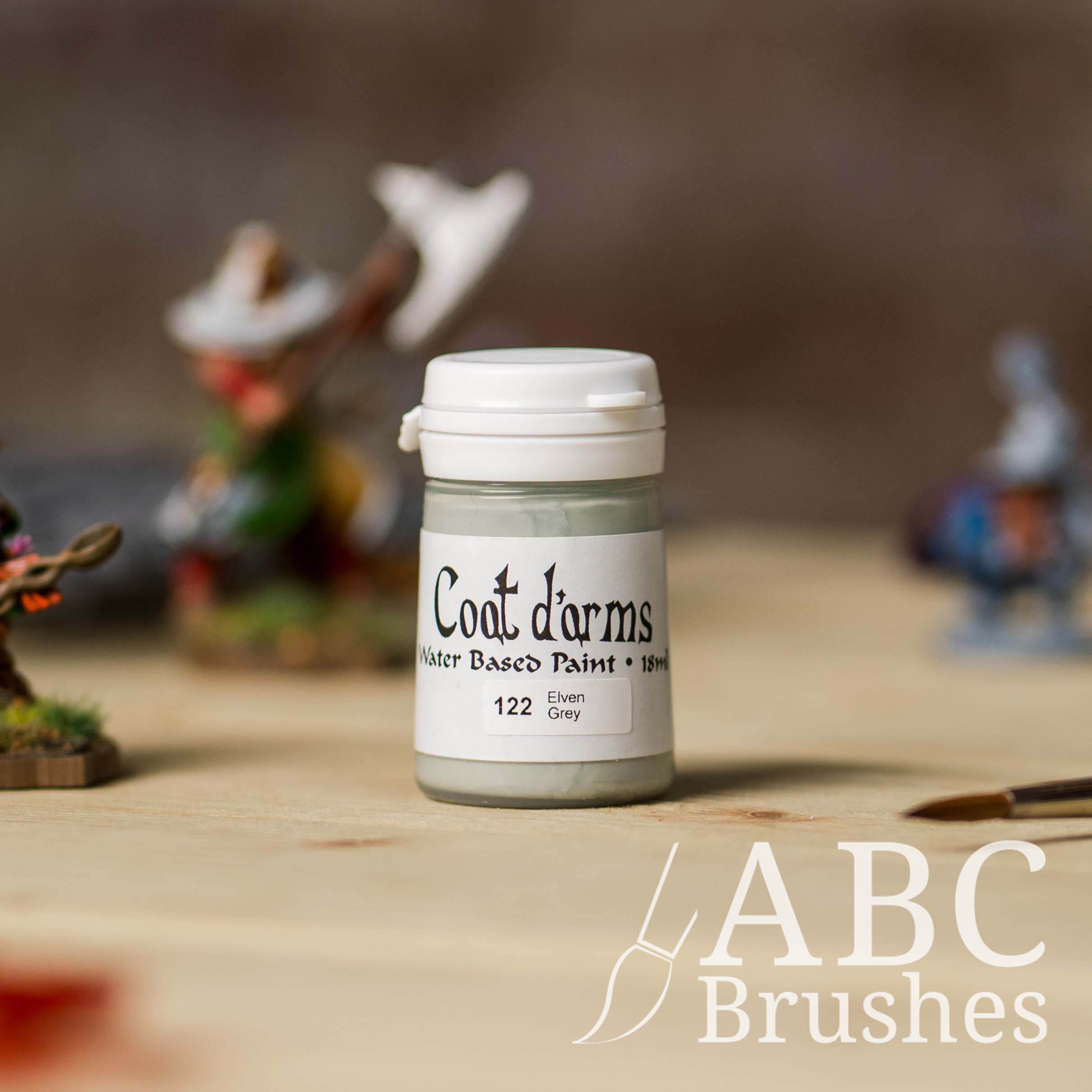 Bottle of Coat d'Arms acrylic paint in the colour Elven Grey, with various wargaming models and an abc paintbrush in the background, on a wooden surface.