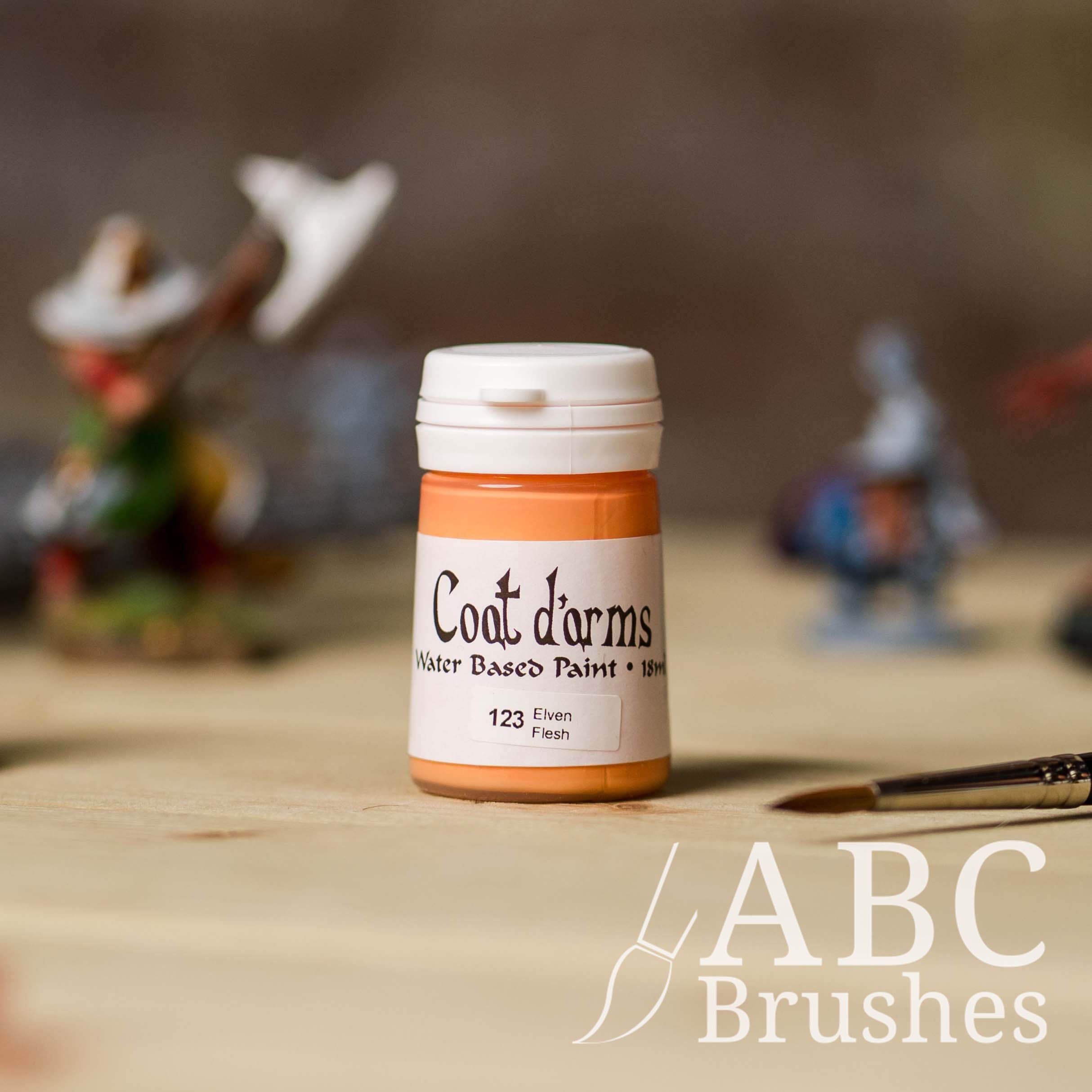 Bottle of Coat d'Arms acrylic paint in the colour Elven Flesh, with various wargaming models and an abc paintbrush in the background, on a wooden surface.