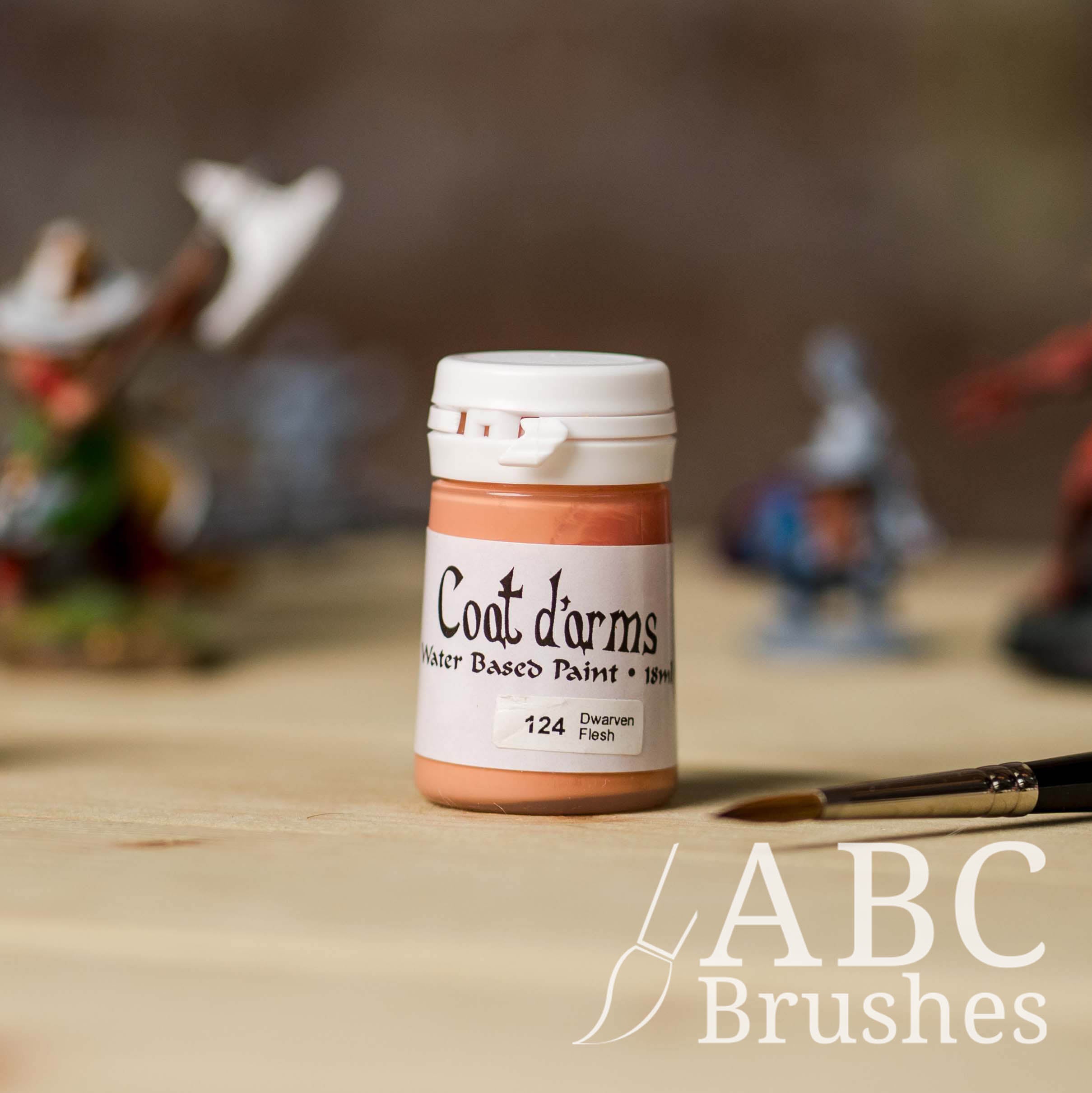 Bottle of Coat d'Arms acrylic paint in the colour Dwarven Flesh, with various wargaming models and an abc paintbrush in the background, on a wooden surface.