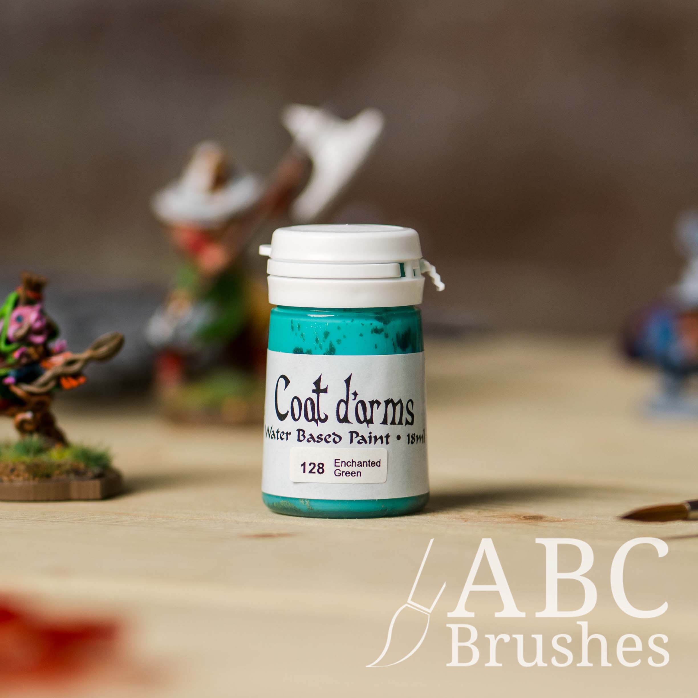Bottle of Coat d'Arms acrylic paint in the colour Enchanted Green, with various wargaming models and an abc paintbrush in the background, on a wooden surface.