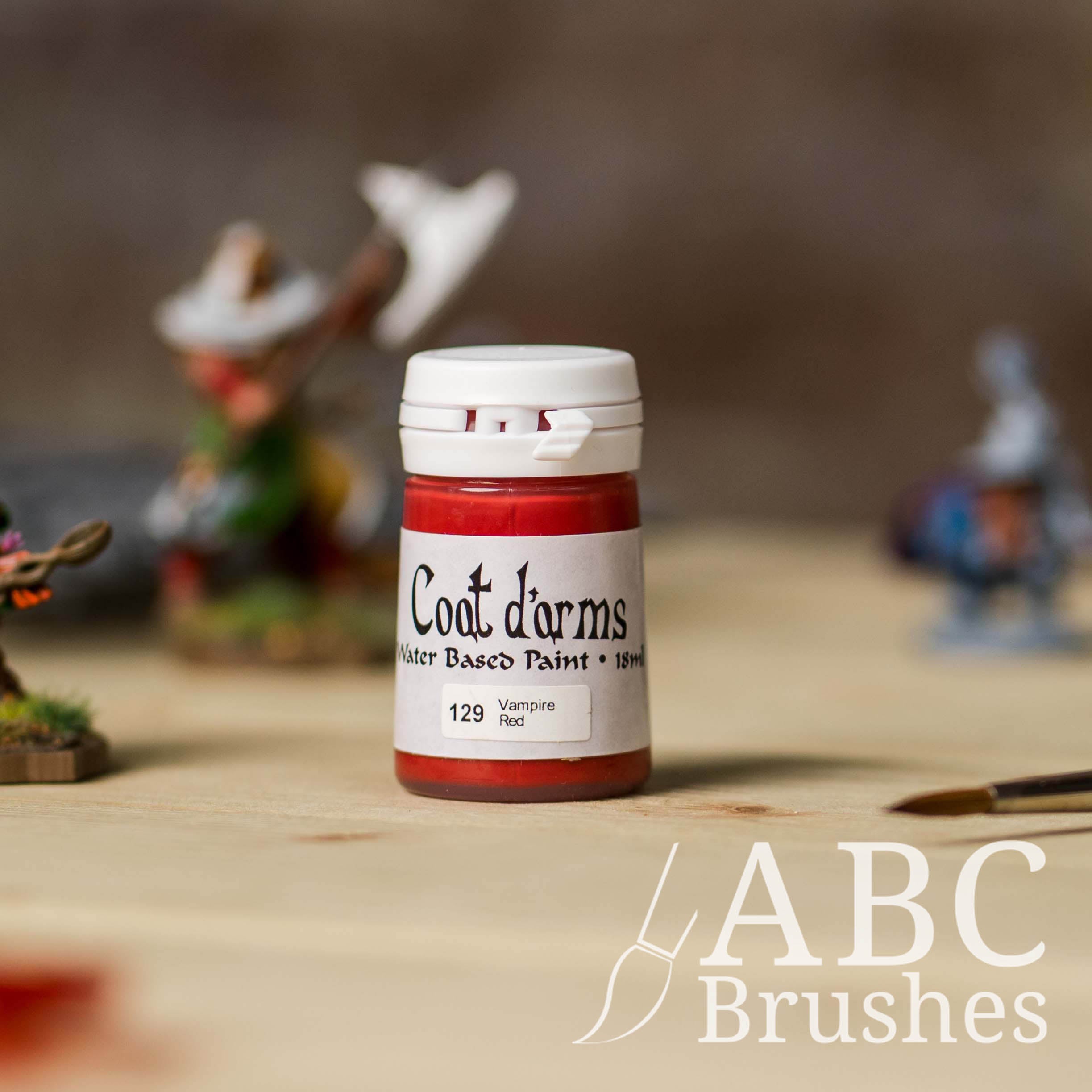 Bottle of Coat d'Arms acrylic paint in the colour Vampire Red, with various wargaming models and an abc paintbrush in the background, on a wooden surface.