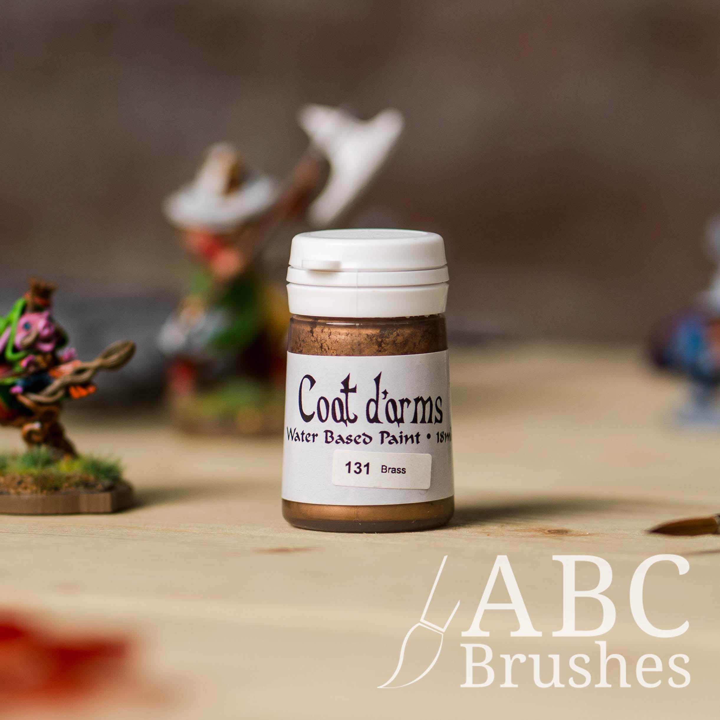 Bottle of Coat d'Arms acrylic paint in the colour Brass, with various wargaming models and an abc paintbrush in the background, on a wooden surface.