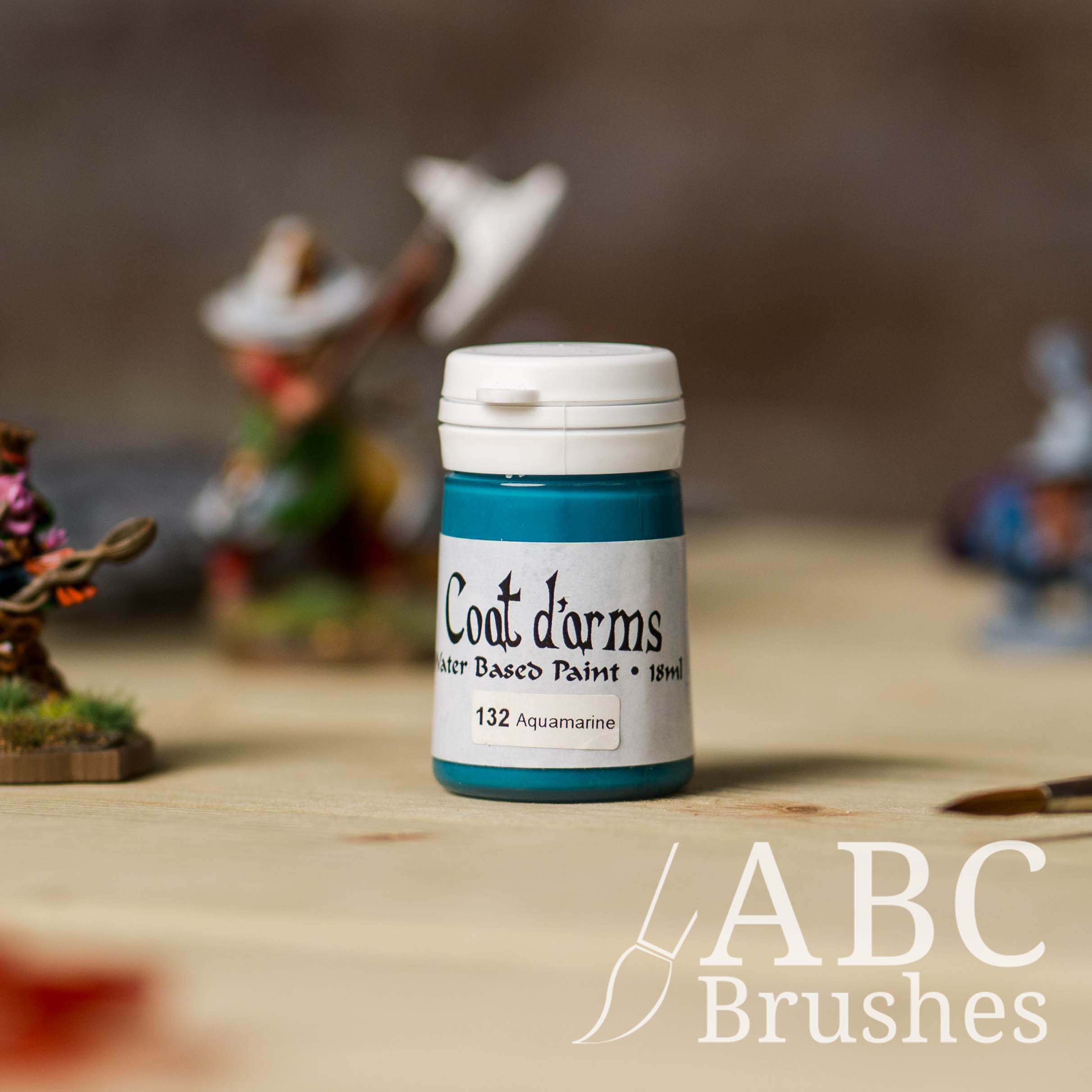 Bottle of Coat d'Arms paint in the colour Aquamarine, with various wargaming models and an abc paintbrush in the background, on a wooden surface.