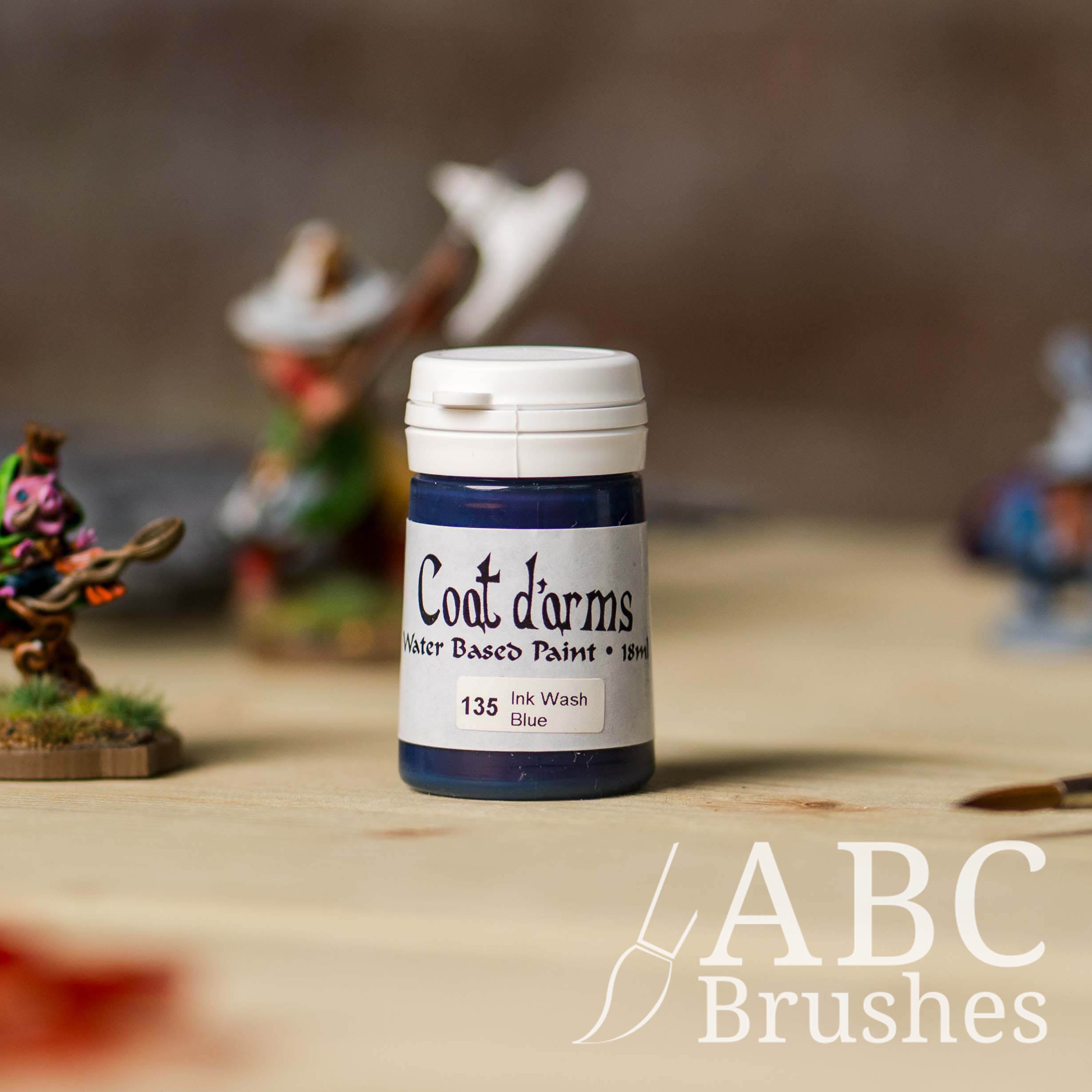 Bottle of Coat d'Arms ink wash in the colour blue, with various wargaming models and an abc paintbrush in the background, on a wooden surface.