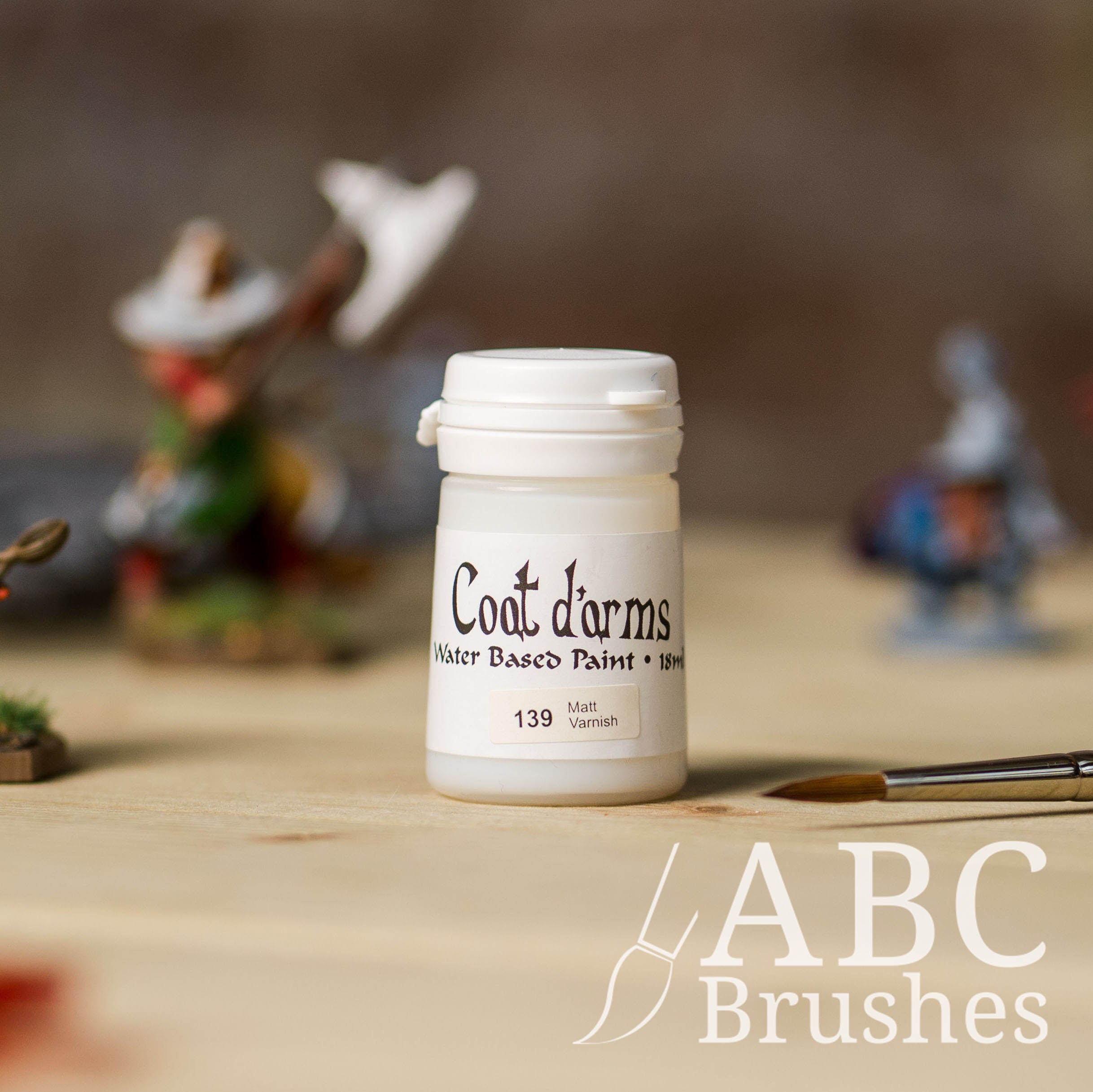 Bottle of Coat d'Arms Matt Varnish, with various wargaming models and an abc paintbrush in the background, on a wooden surface.