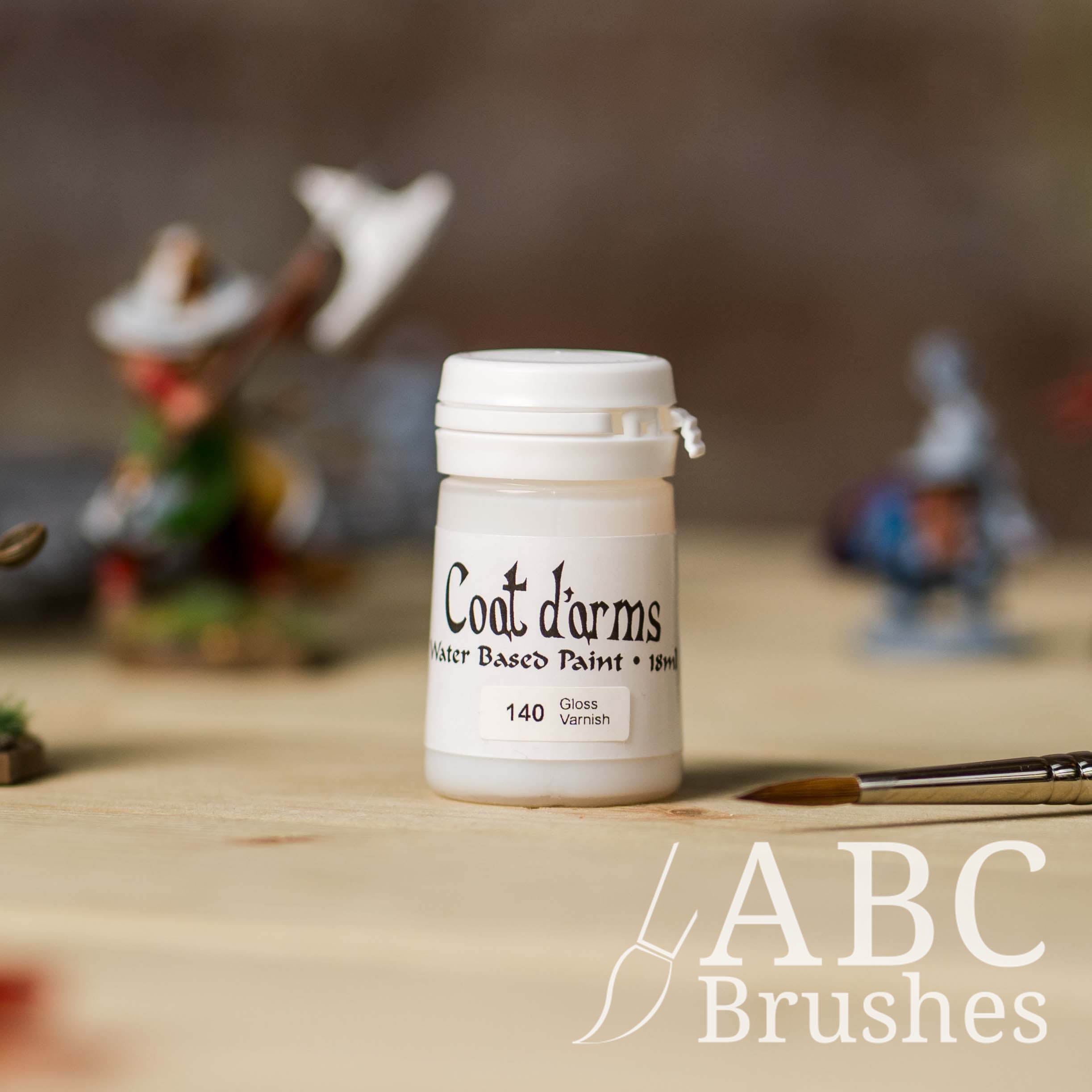 Bottle of Coat d'Arms Gloss Varnish, with various wargaming models and an abc paintbrush in the background, on a wooden surface.