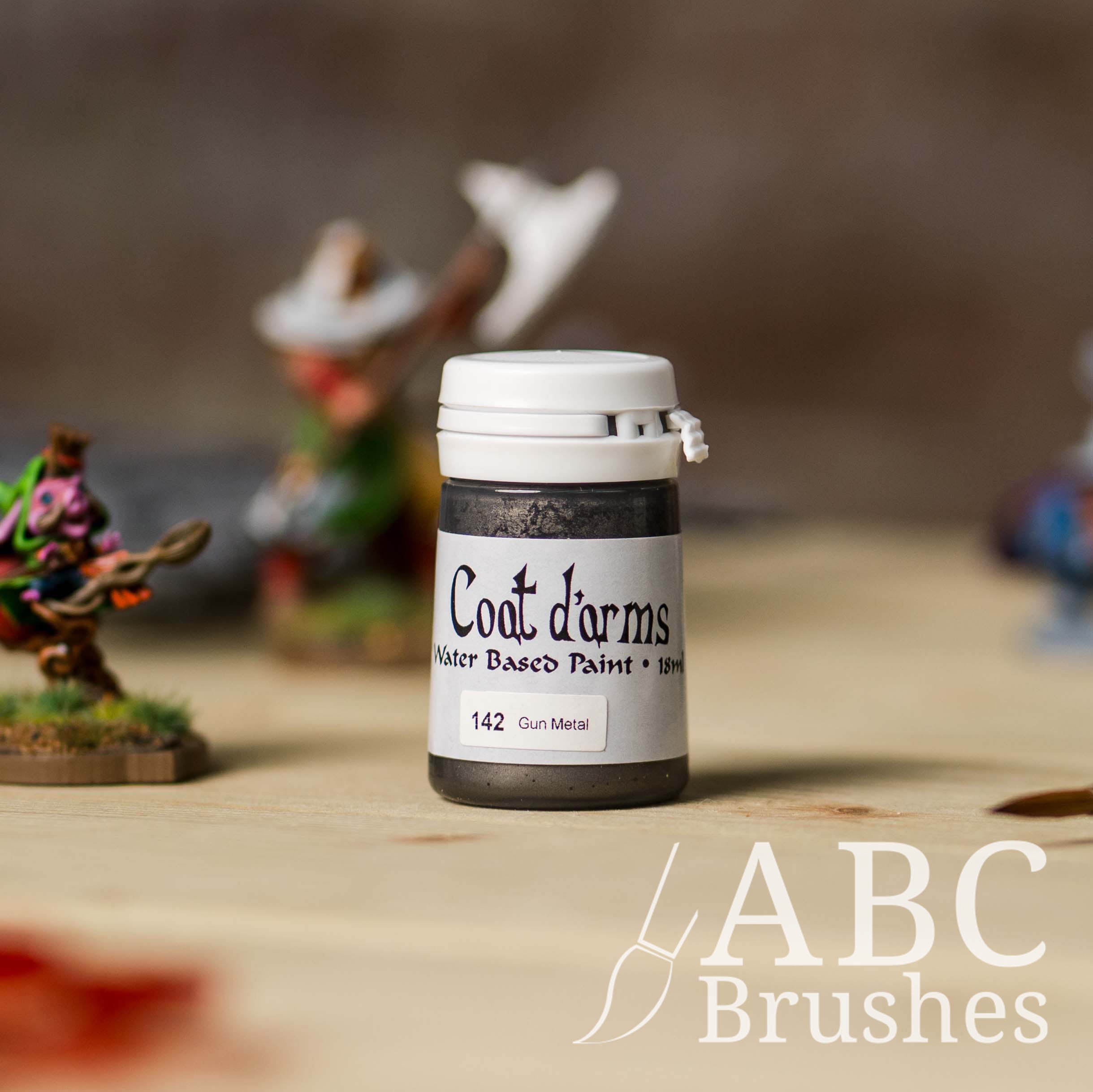 Bottle of Coat d'Arms paint in the colour Gun Metal, with various wargaming models and an abc paintbrush in the background, on a wooden surface.