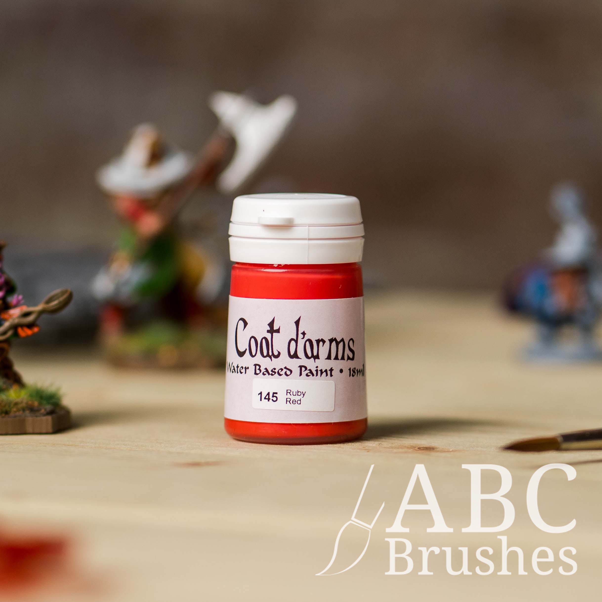 Bottle of Coat d'Arms paint in the colour Ruby Red, with various wargaming models and an abc paintbrush in the background, on a wooden surface.