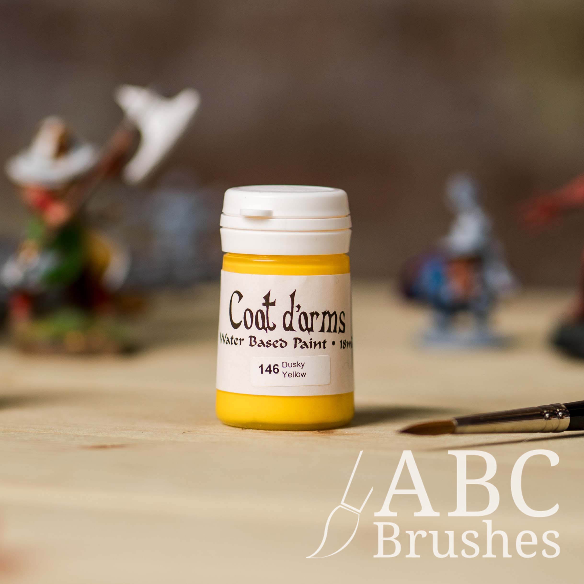 Bottle of Coat d'Arms paint in the colour Dusky Yellow, with various wargaming models and an abc paintbrush in the background, on a wooden surface.