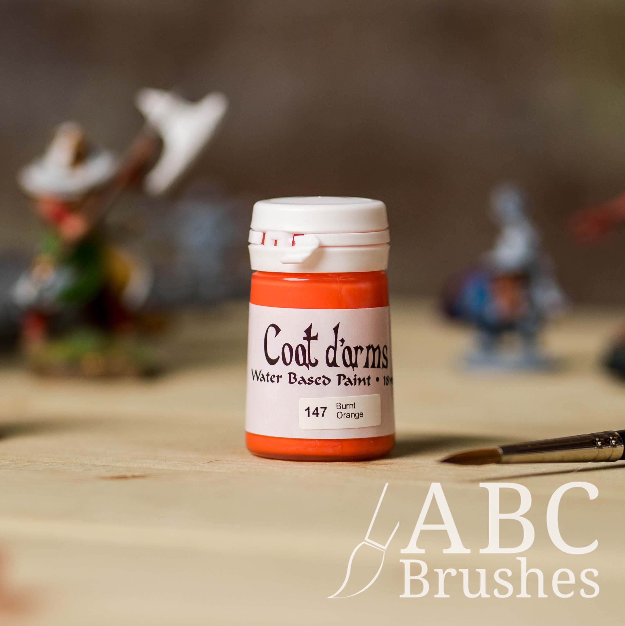 Bottle of Coat d'Arms paint in the colour Burnt Orange, with various wargaming models and an abc paintbrush in the background, on a wooden surface.
