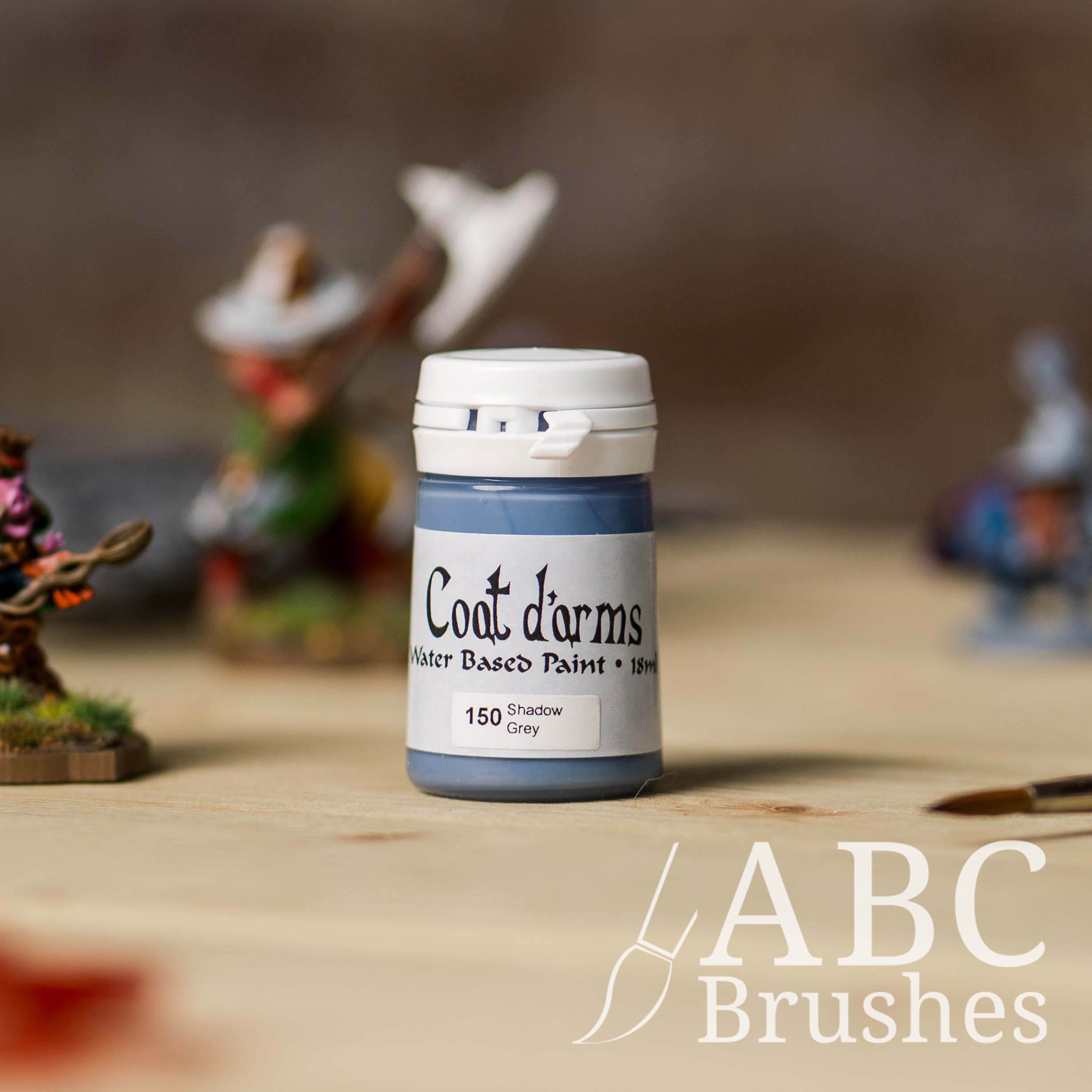 Bottle of Coat d'Arms paint in the colour Shadow Grey, with various wargaming models and an abc paintbrush in the background, on a wooden surface.