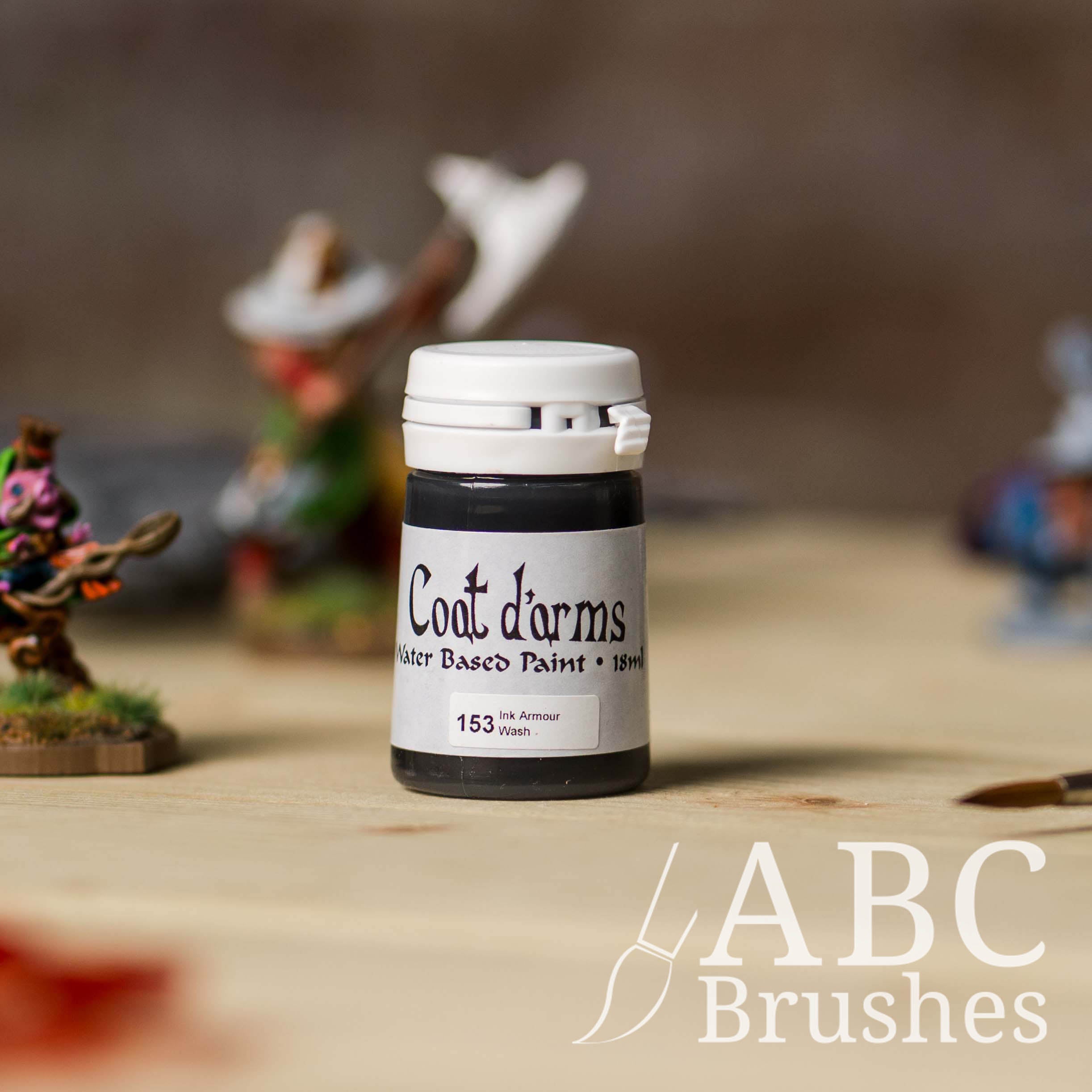 Bottle of Coat d'Arms paint in the colour Ink Armour Wash, with various wargaming models and an abc paintbrush in the background, on a wooden surface.