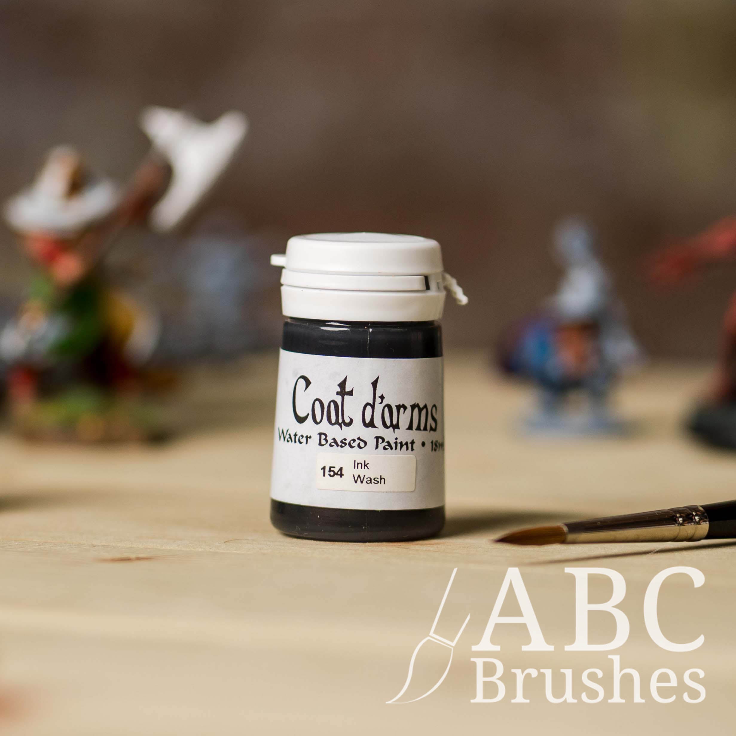 Bottle of Coat d'Arms paint in the colour Ink wash, with various wargaming models and an abc paintbrush in the background, on a wooden surface.