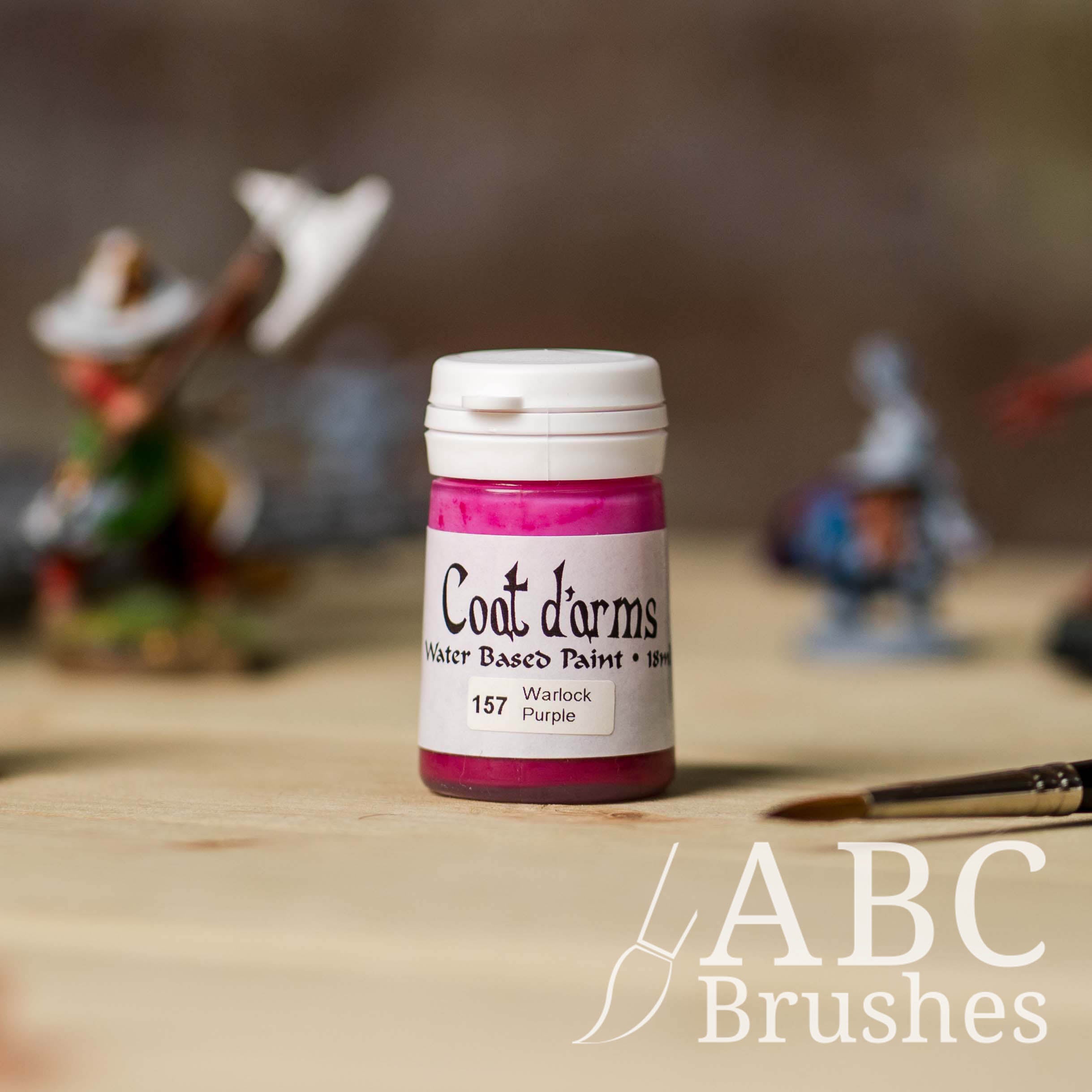 Bottle of Coat d'Arms Acrylic paint in the colour Warlock Purple, with various wargaming models and an abc paintbrush in the background, on a wooden surface.