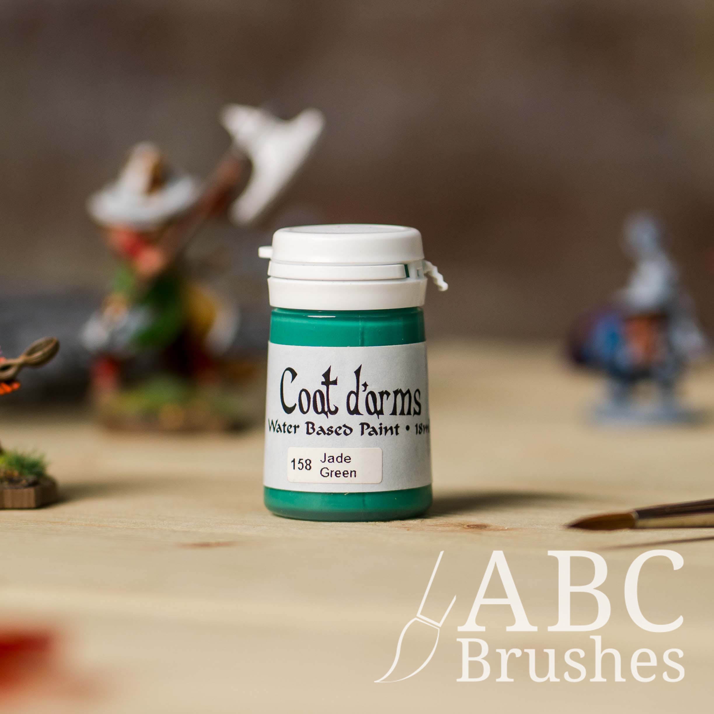 Bottle of Coat d'Arms Acrylic paint in the colour Jade Green, with various wargaming models and an abc paintbrush in the background, on a wooden surface.