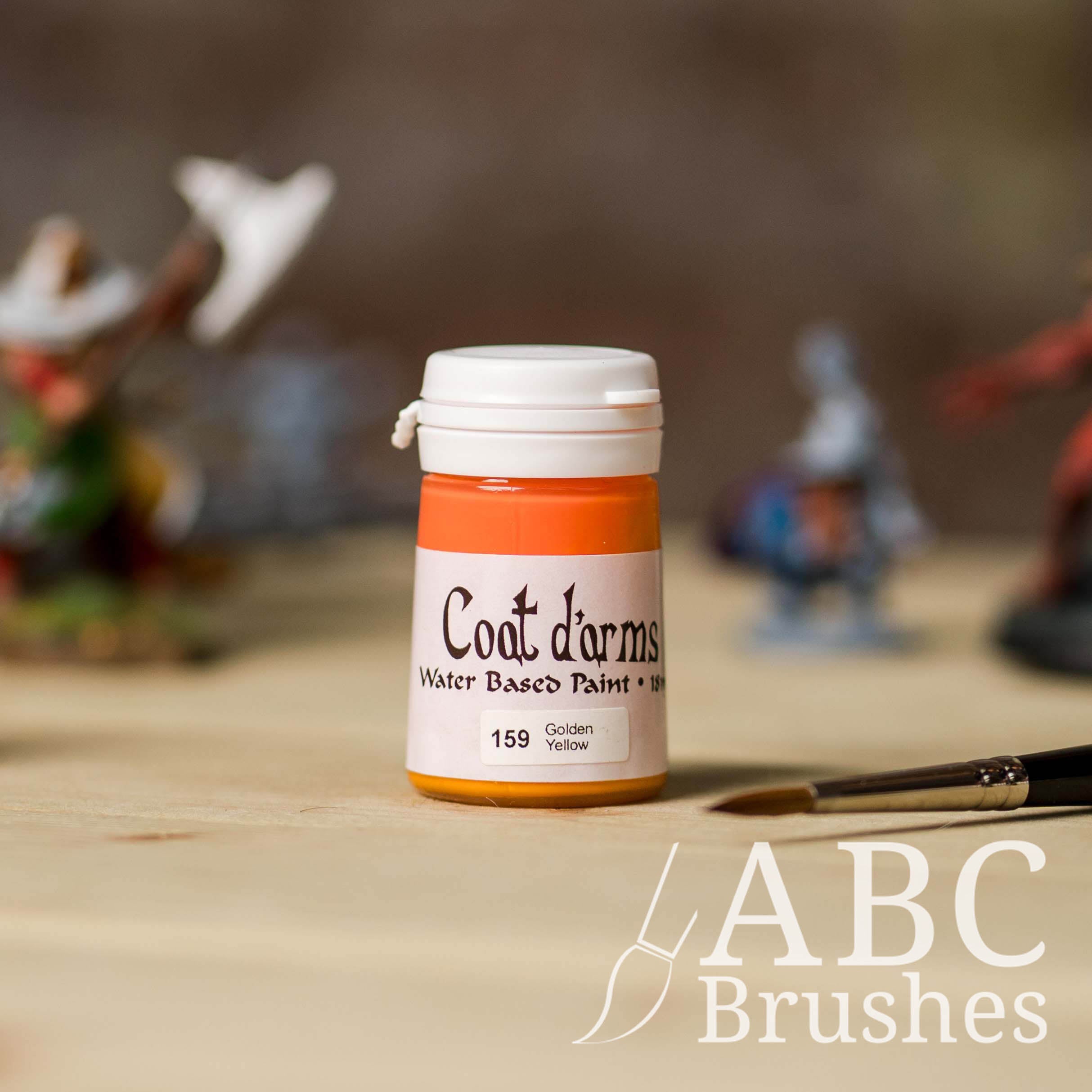 Bottle of Coat d'Arms Acrylic paint in the colour Golden Yellow, with various wargaming models and an abc paintbrush in the background, on a wooden surface.