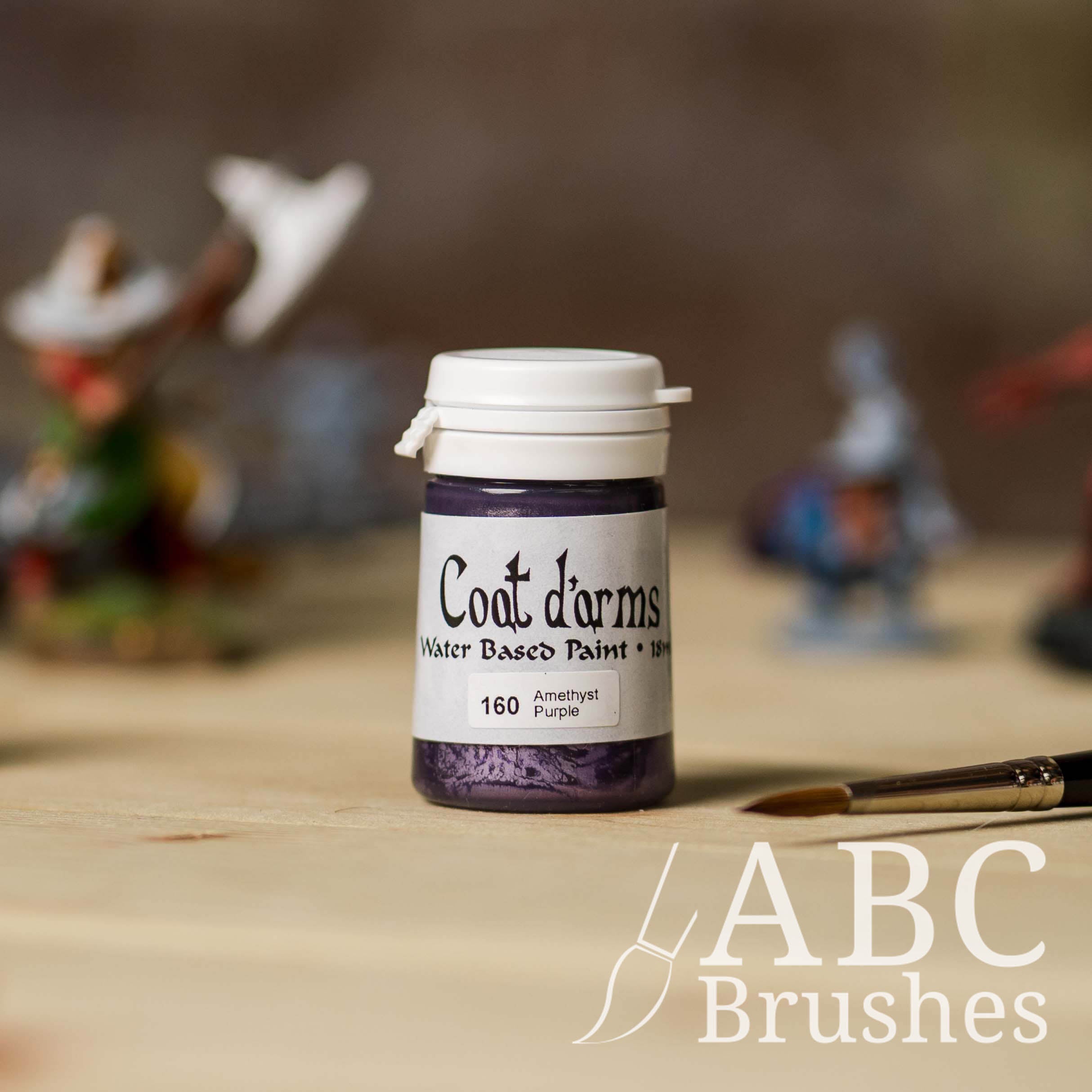 Bottle of Coat d'Arms Acrylic paint in the colour Amethyst Purple, with various wargaming models and an abc paintbrush in the background, on a wooden surface.