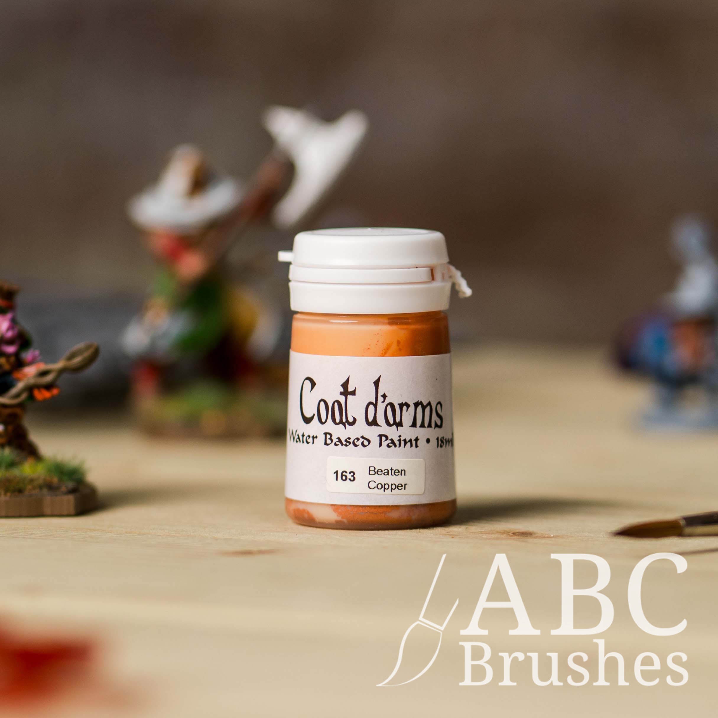 Bottle of Coat d'Arms Acrylic paint in the colour Beaten Copper, with various wargaming models and an abc paintbrush in the background, on a wooden surface.