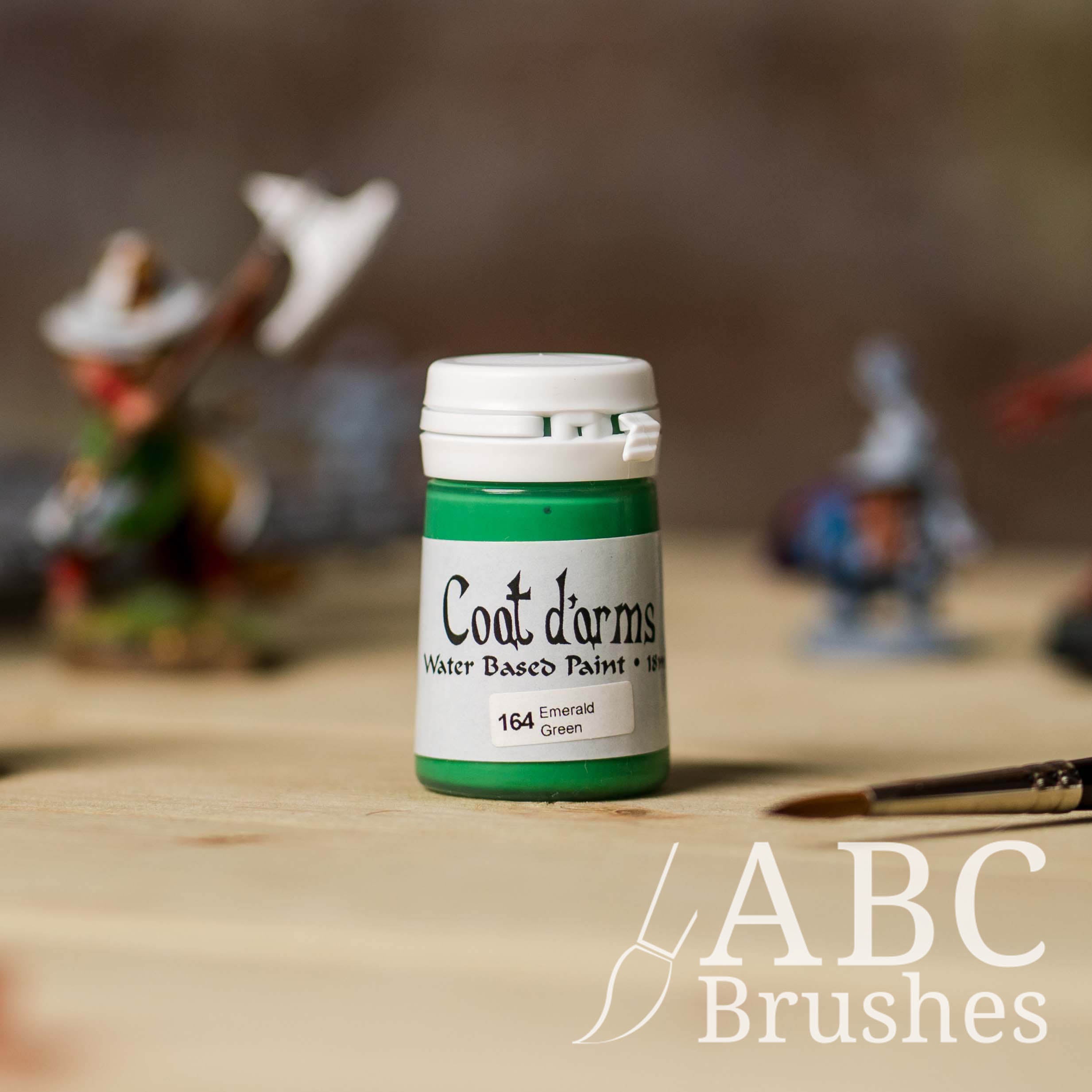 Bottle of Coat d'Arms Acrylic paint in the colour Emerald Green, with various wargaming models and an abc paintbrush in the background, on a wooden surface.
