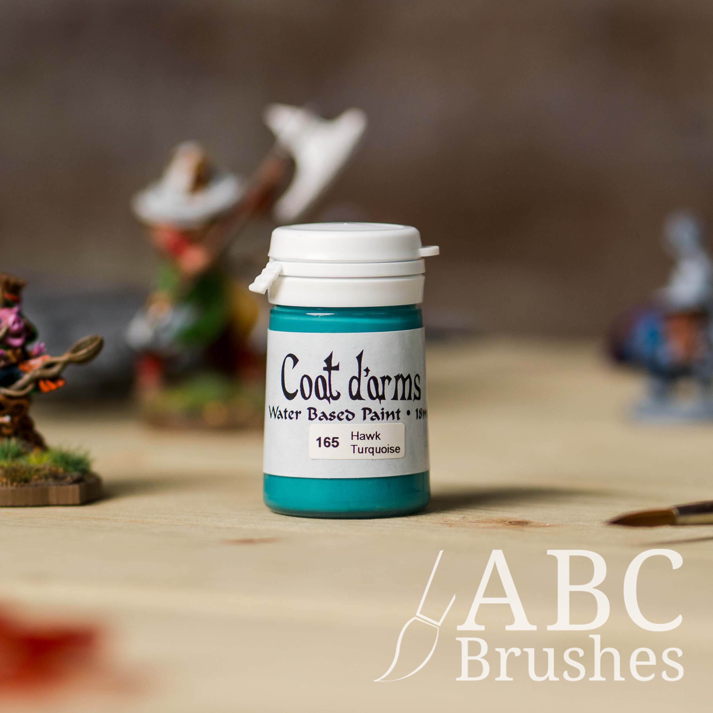 Bottle of Coat d'Arms Acrylic paint in the colour Hawk Turquoise, with various wargaming models and an abc paintbrush in the background, on a wooden surface.