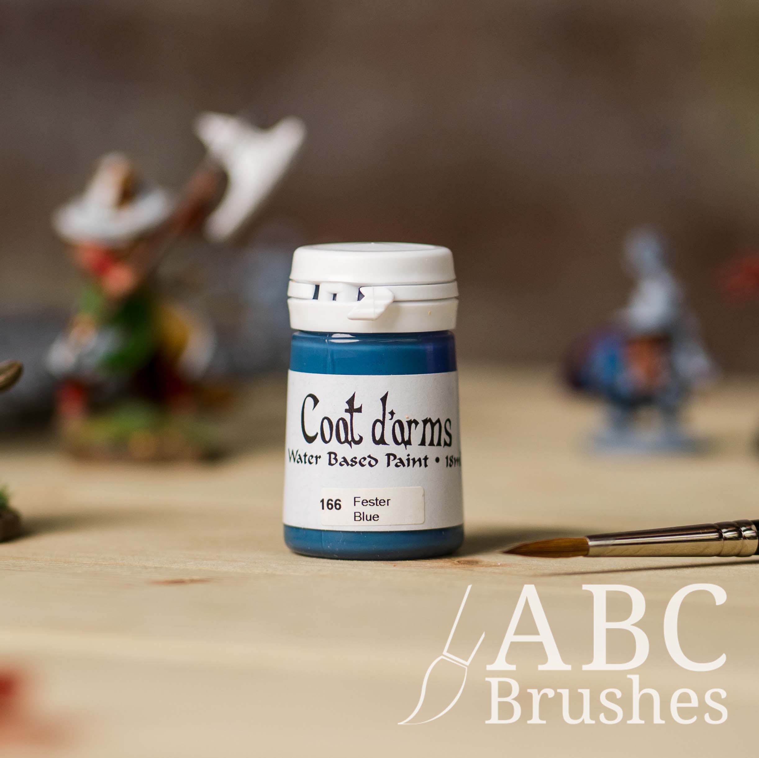 Bottle of Coat d'Arms Acrylic paint in the colour Fester Blue, with various wargaming models and an abc paintbrush in the background, on a wooden surface.