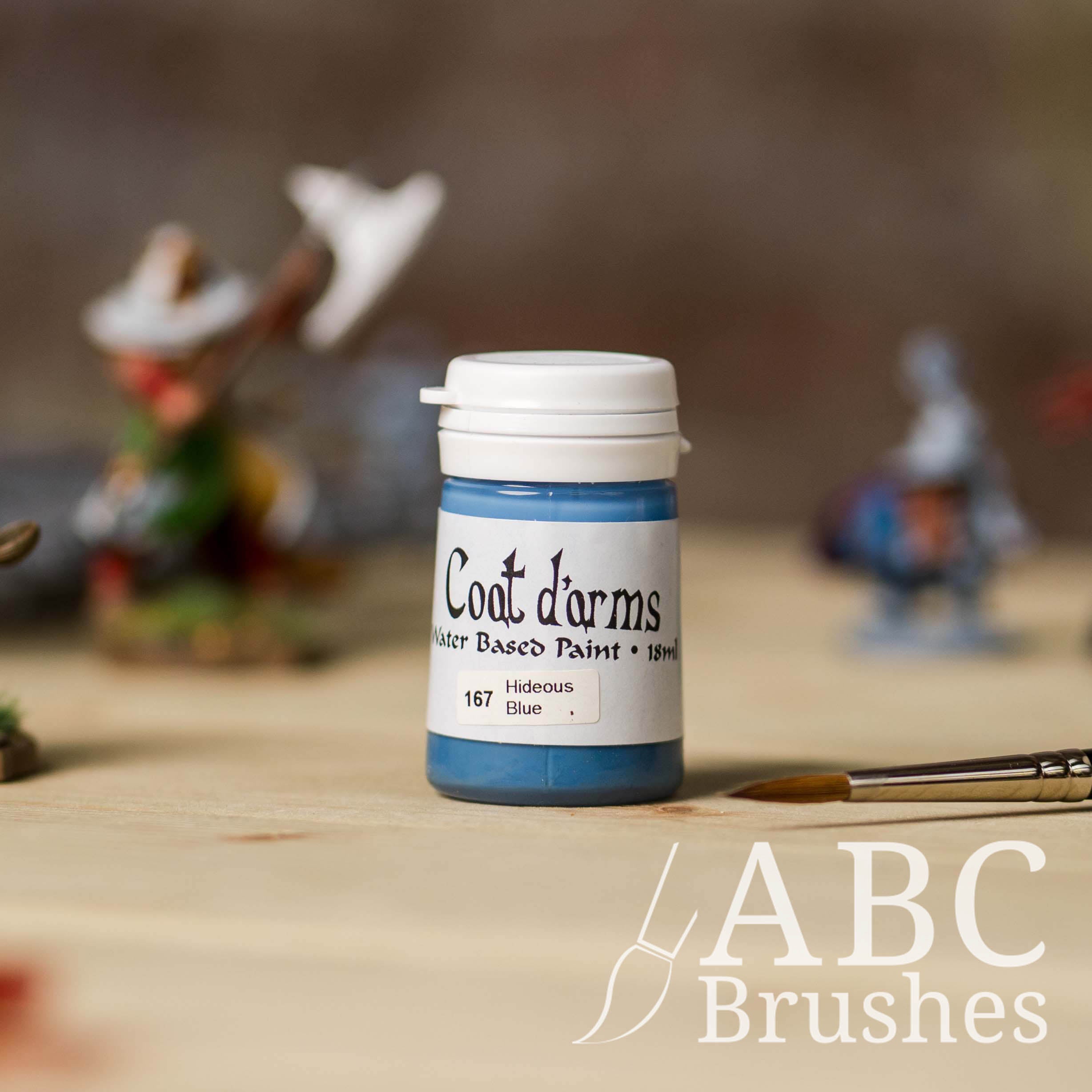 Bottle of Coat d'Arms Acrylic paint in the colour Hideous Blue, with various wargaming models and an abc paintbrush in the background, on a wooden surface.