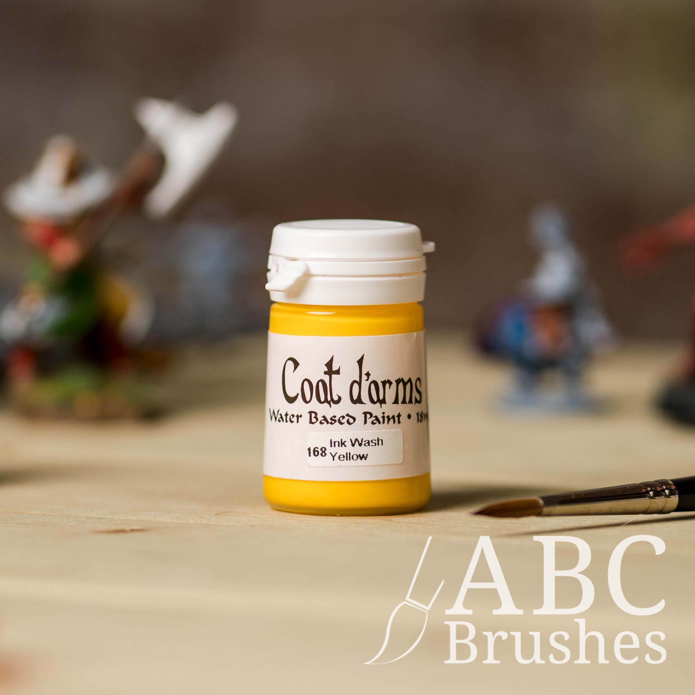 Bottle of Coat d'Arms Ink Wash paint in the colour Yellow, with various wargaming models and an abc paintbrush in the background, on a wooden surface.