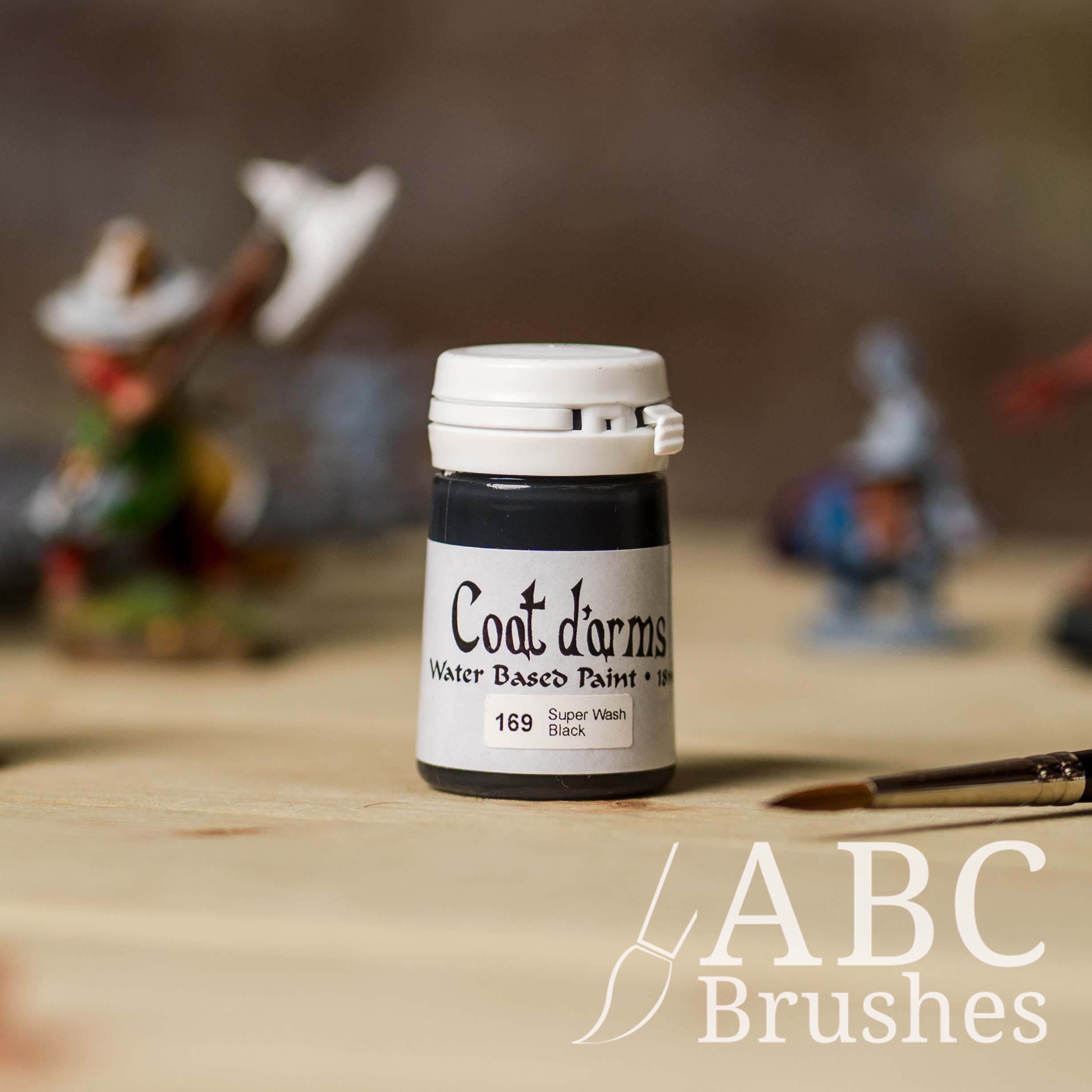 Bottle of Coat d'Arms Super Wash paint in the colour Black with various wargaming models and an abc paintbrush in the background, on a wooden surface.