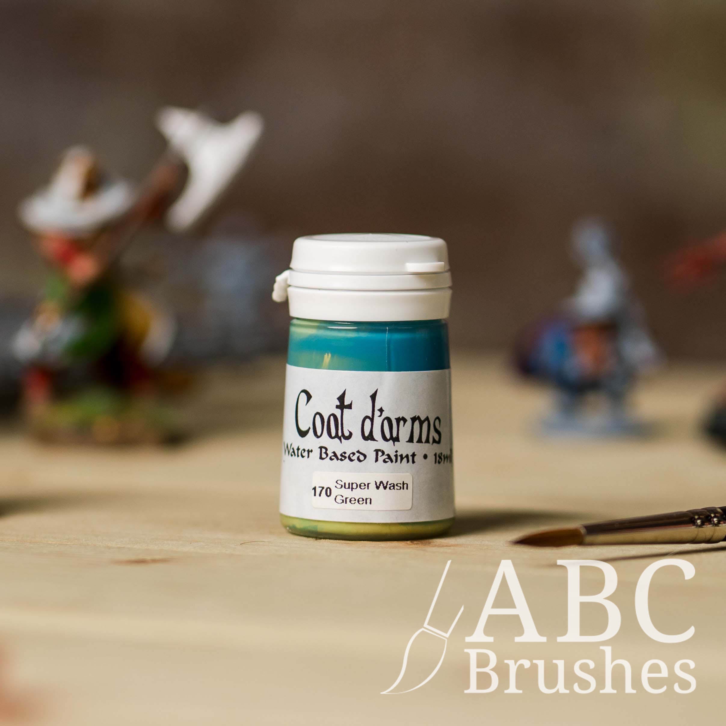 Bottle of Coat d'Arms Super Wash paint in the colour Green, with various wargaming models and an abc paintbrush in the background, on a wooden surface.