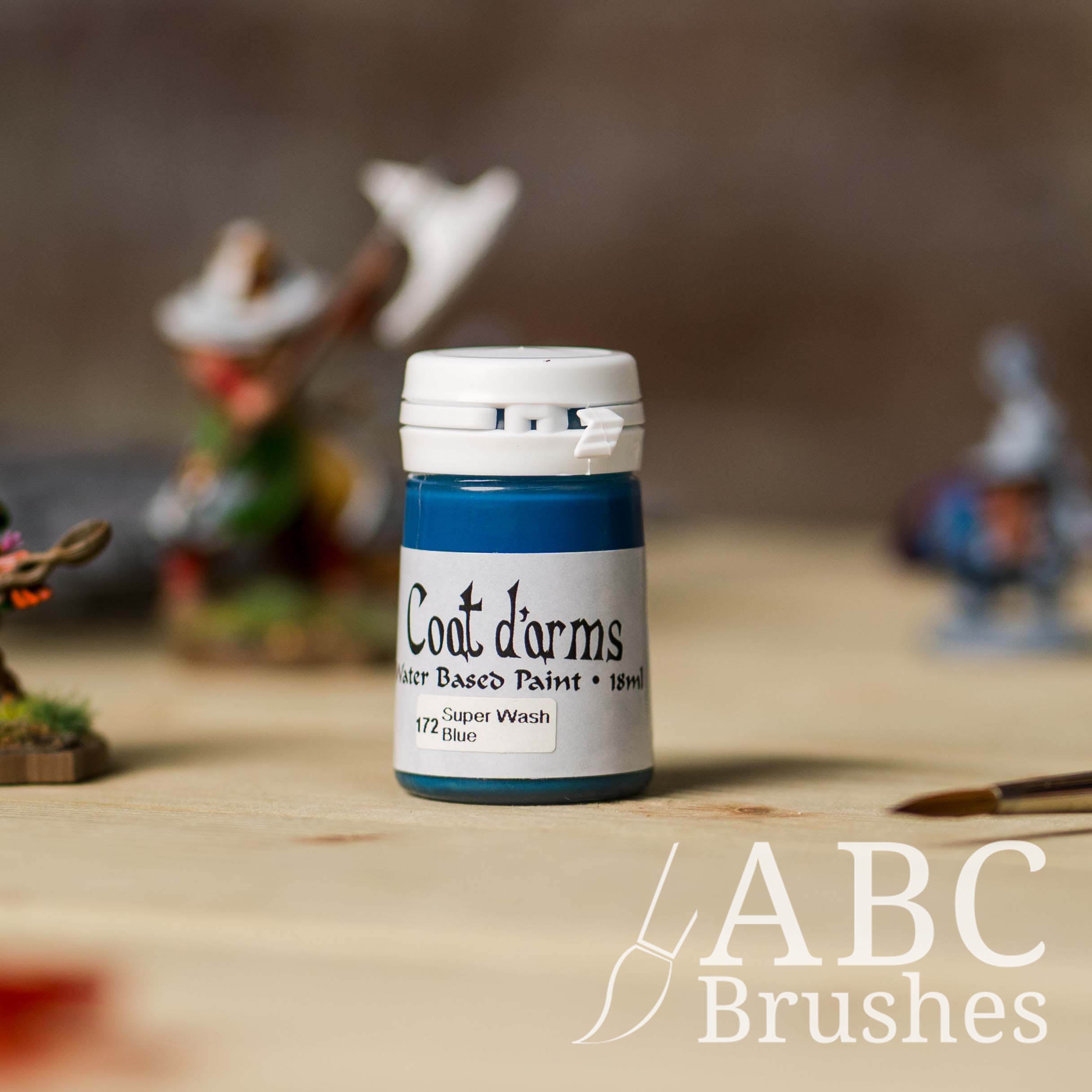 Bottle of Coat d'Arms Super Wash paint in the colour Blue, with various wargaming models and an abc paintbrush in the background, on a wooden surface.