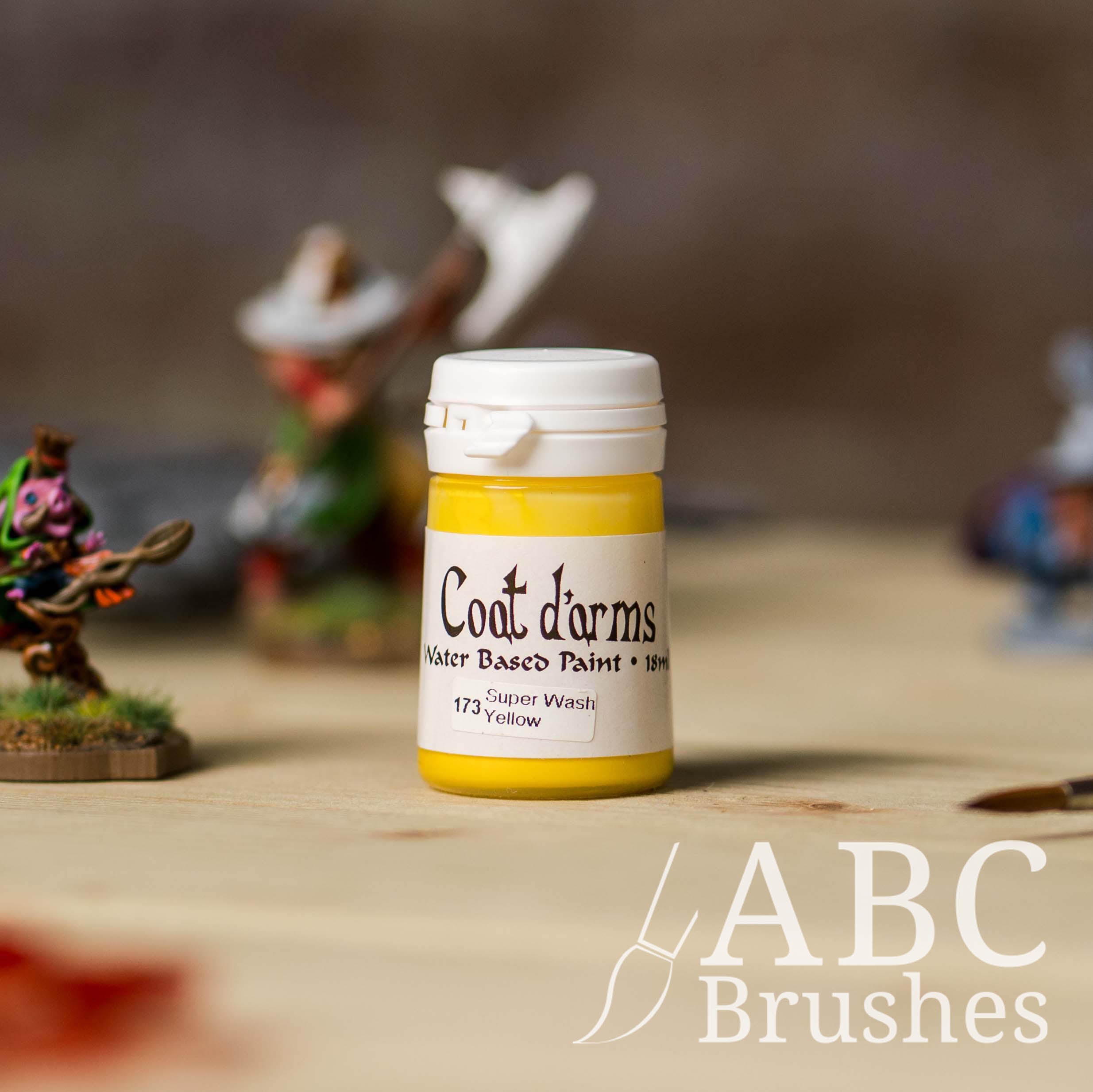 Bottle of Coat d'Arms Super Wash paint in the colour Yellow, with various wargaming models and an abc paintbrush in the background, on a wooden surface.