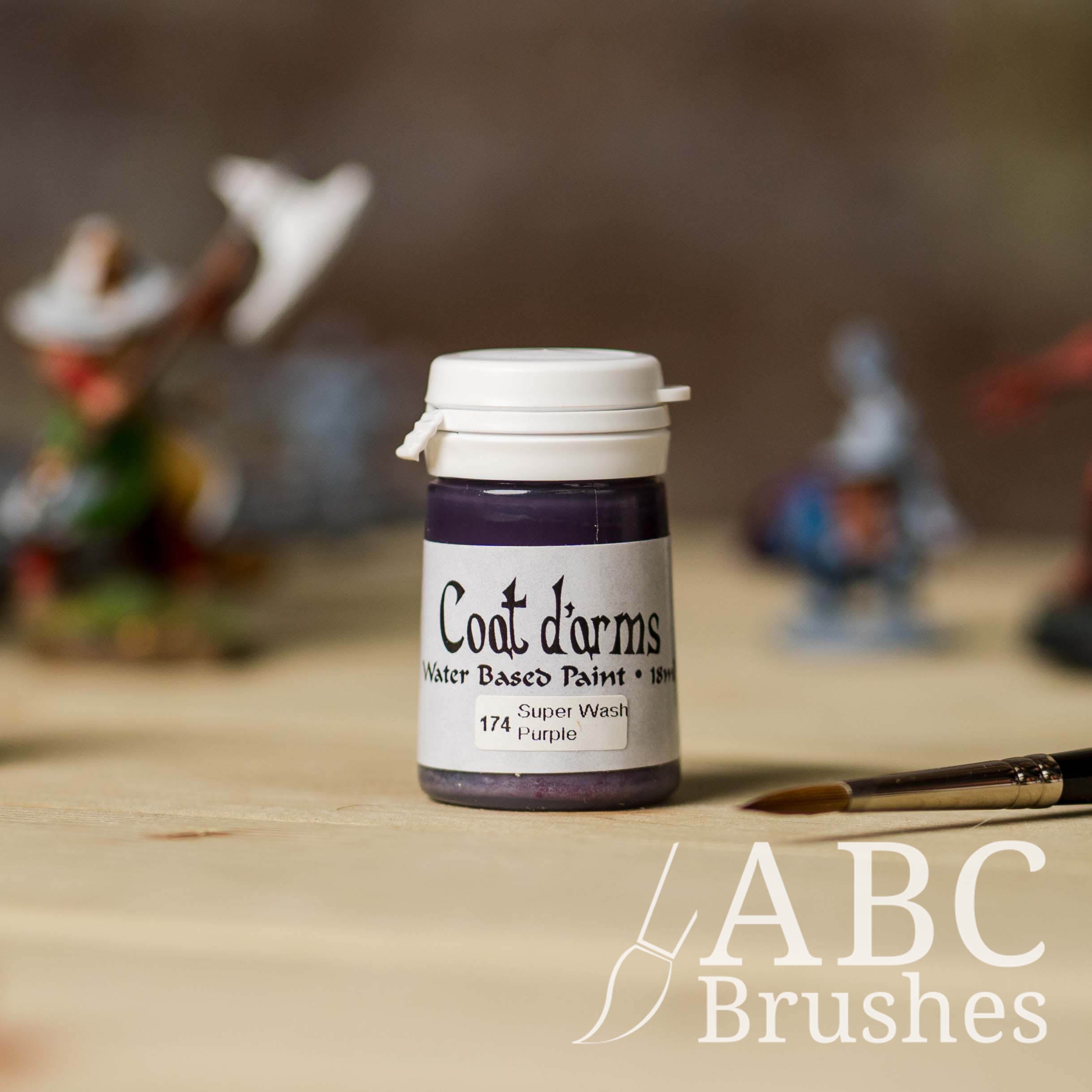 Bottle of Coat d'Arms Super Wash paint in the colour Purple, with various wargaming models and an abc paintbrush in the background, on a wooden surface.