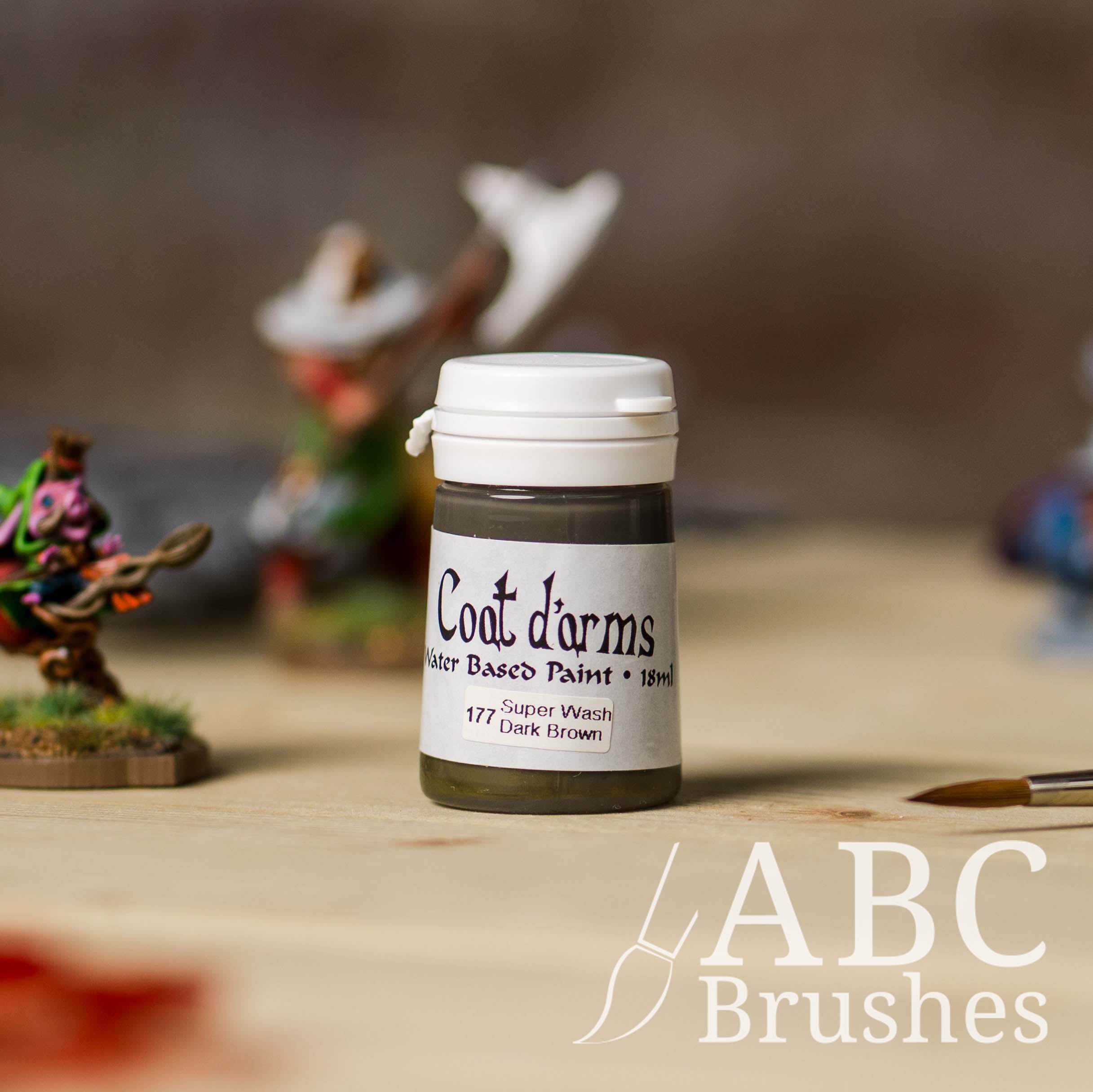 Bottle of Coat d'Arms Super Wash paint in the colour Dark Brown, with various wargaming models and an abc paintbrush in the background, on a wooden surface.