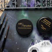 Two ABC Brushes brush cleaners placed on a paint covered cutting mat with various miniature painting supplies placed around them. 