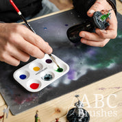 Someone picking up paint from an ABC Brushes paint palette, to apply to a miniature in their other hand. 
