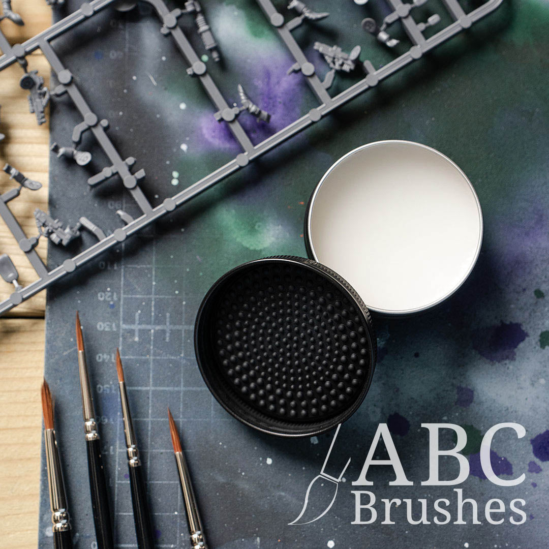 Open ABC Brushes brush cleaner, with the textured lid next to it, placed on a paint covered cutting mat with various miniature painting supplies placed around it. 