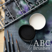 Open ABC Brushes brush cleaner, with the textured lid next to it, placed on a paint covered cutting mat with various miniature painting supplies placed around it. 