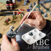 Using a Pro Arte White Nylon flat paint brush to paint the base of a miniature in dark blue paint, with various mini figures and painting supplies in the background. 