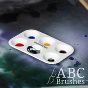 ABC Brushes paint palette filled with various coloured acrylic paints, on a paint covered background.