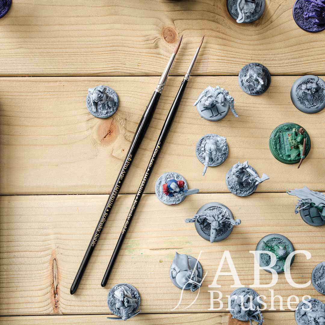 Two Pro Arte Imperium paint brushes on a wooden background surrounded by various painted and unpainted mini figures.