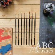 Pro Arte Imperium paint brush range on a wooden background and next to a cutting mat, dry brush board, acrylic paint and a mini on a painting handle.