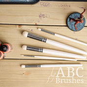ABC Brushes Dry Brush set on a wooden background, next to acrylic paint bottles and a miniature. 